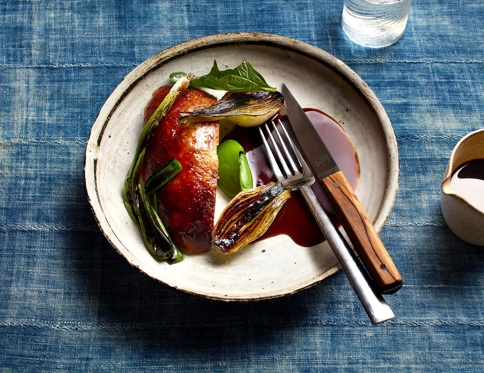 roasted chicken with charred scallions, green beans and dark sauce on a cream plate, set on blue indigo cloth with a knife and fork