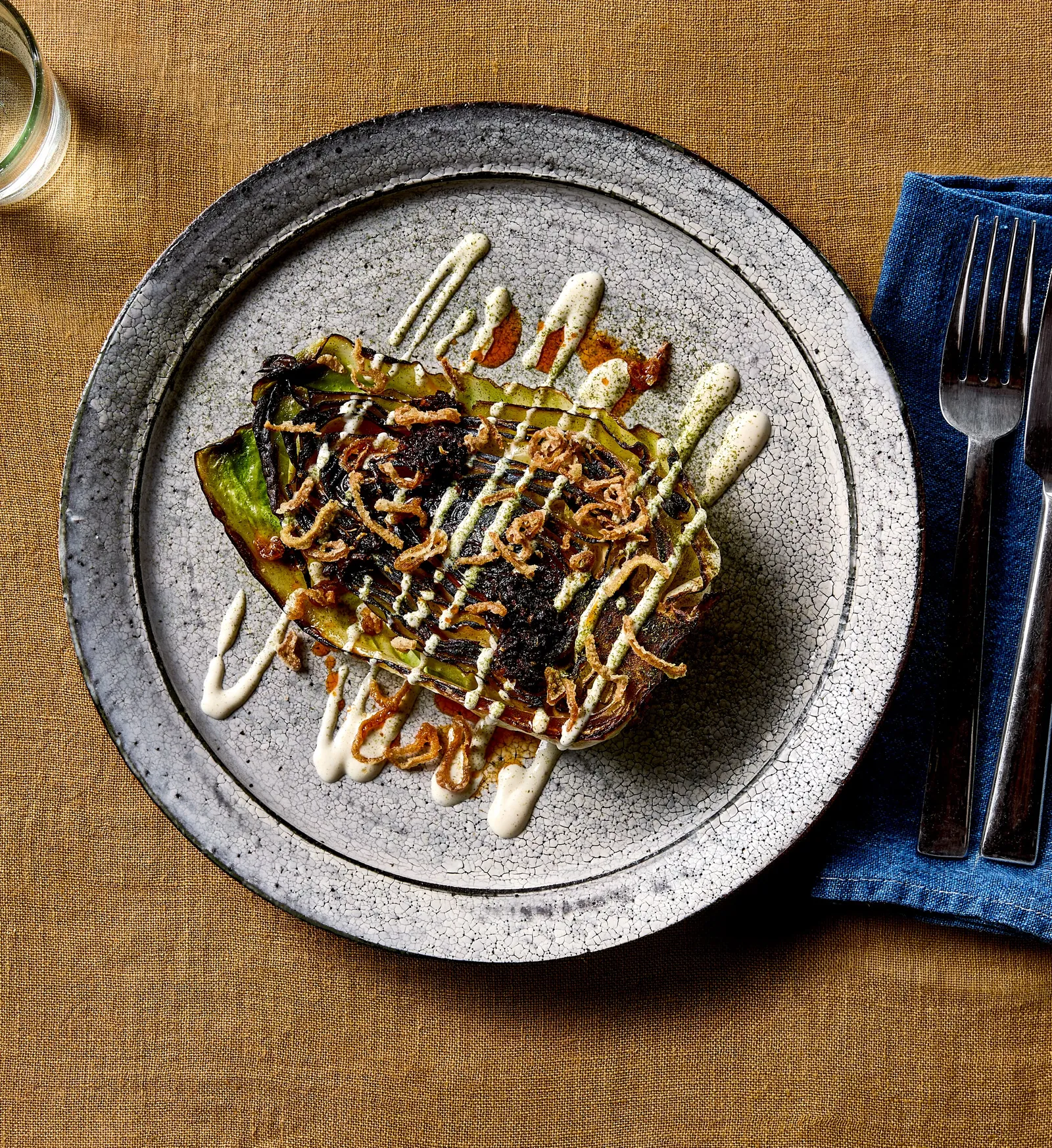 vertical overhead view of charred cabbage with dotted cream sauce and crispy shallots on a crackled gray plate, on mustard linen with a blue napkin