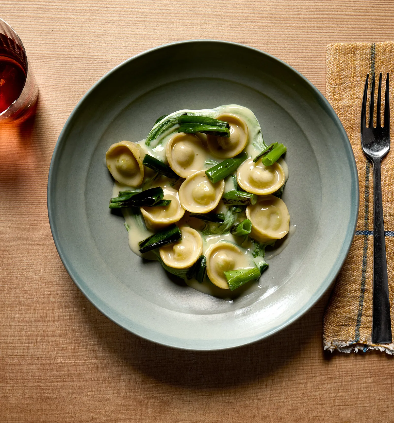 vertical overhead view of small stuffed pasta in pale green sauce with scallions on a blue-gray plate, on light wood with a checked napkin