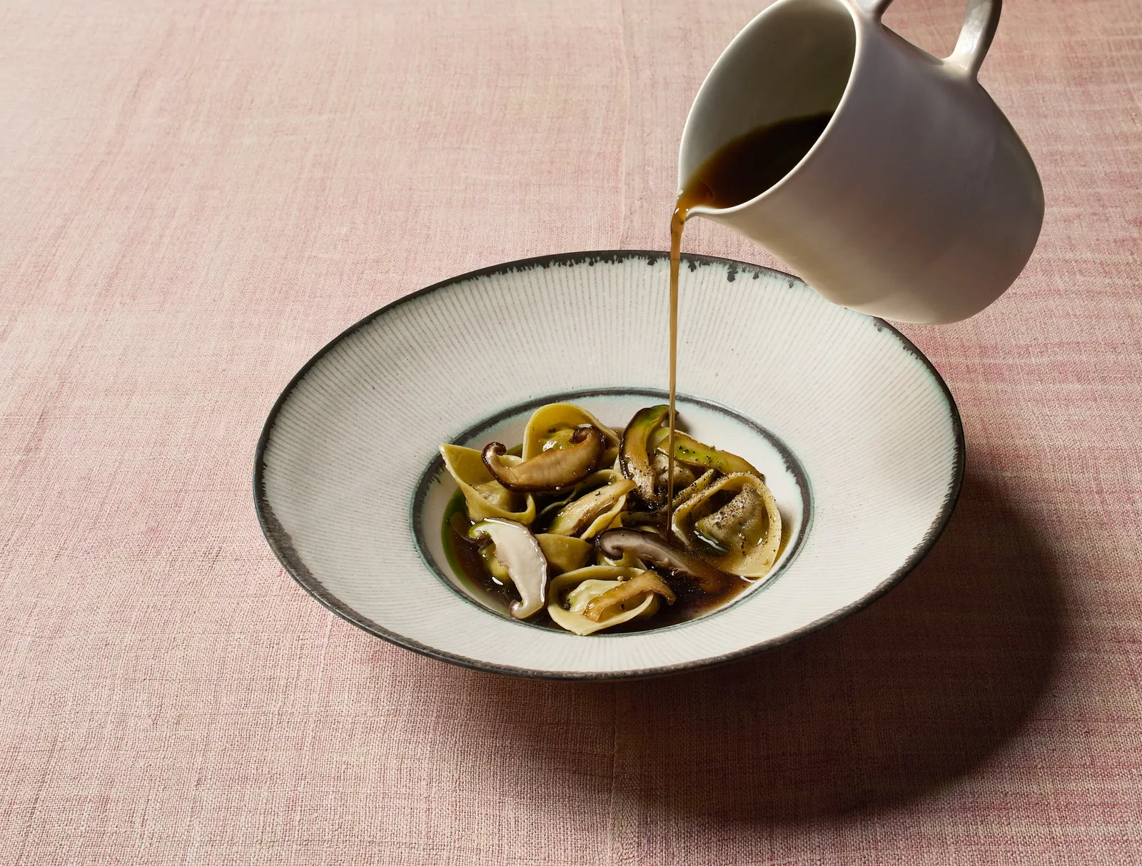 broth poured from a white pitcher over tortellini and mushrooms in a wide striped bowl, on pink linen