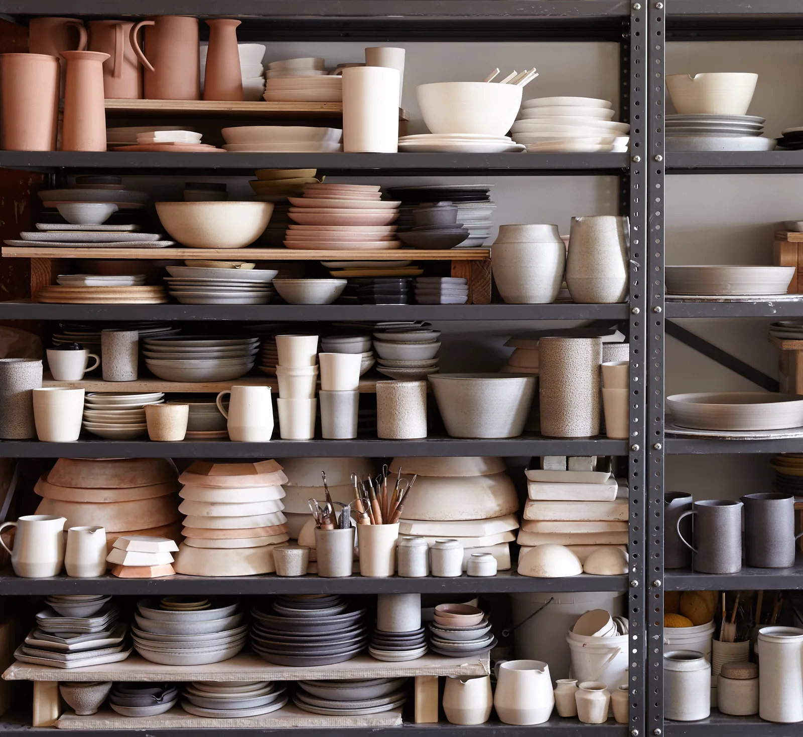 dark metal shelving packed with stoneware: pink clay pitchers, stacked plates, mugs, bowls and brushes in cups, in pinks, greys and creams