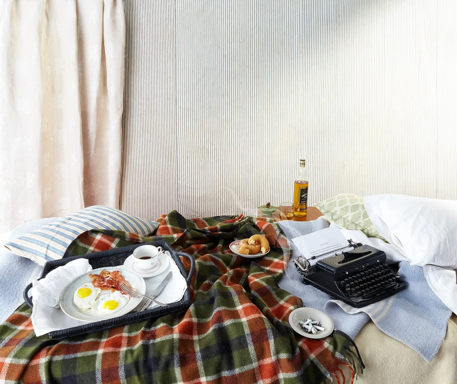 bed under a plaid blanket with a tray of fried eggs, bacon and coffee, a typewriter, a bottle, snails on a saucer and pillows, against a striped wall