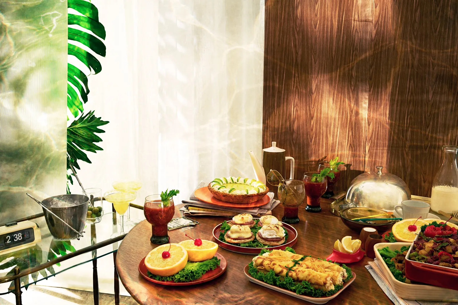 close view of a round wood table with grapefruit halves, eggs, crepes, cocktails, a pie and a covered dish, a sunlit window and palm at left