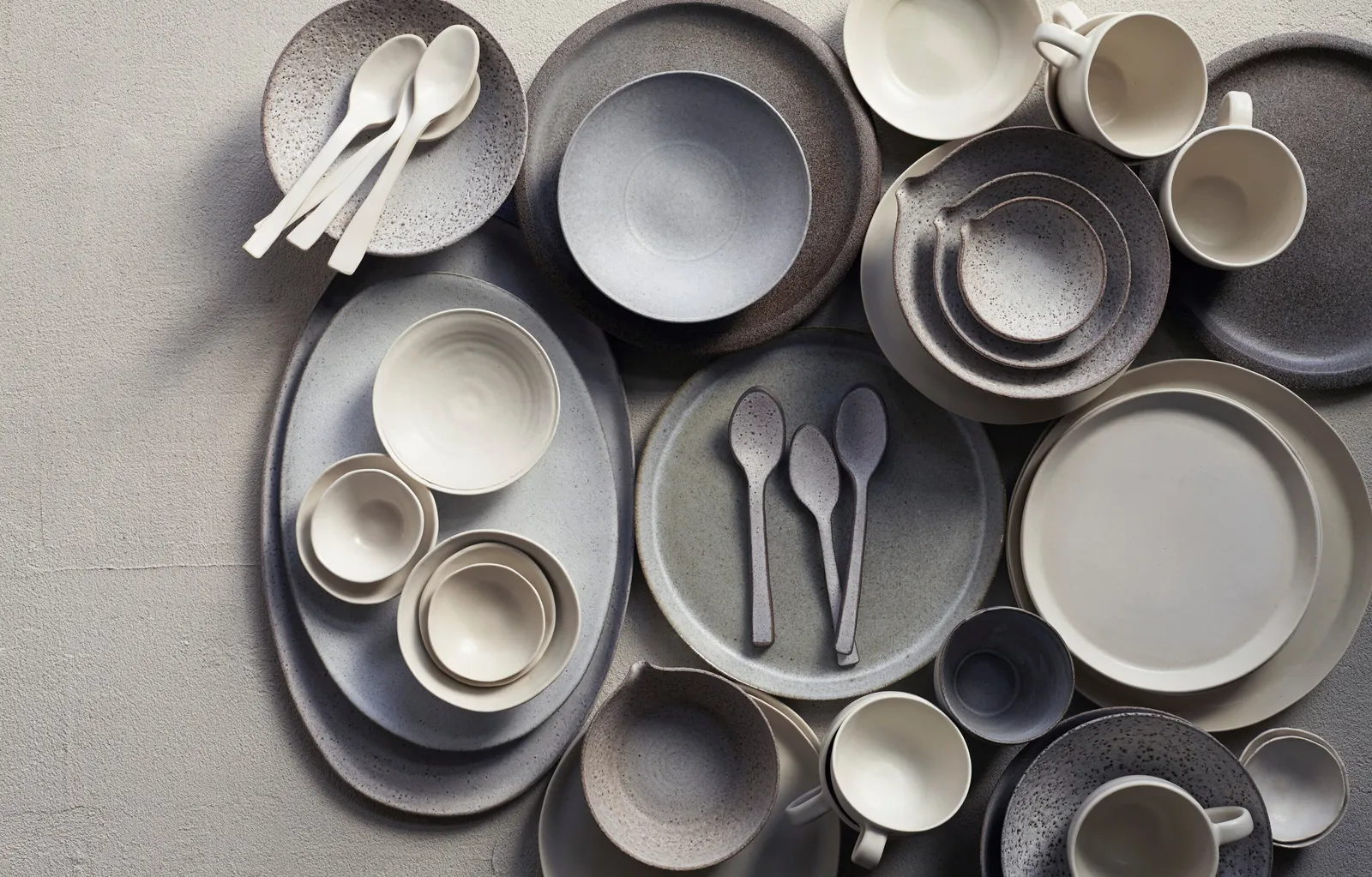 overhead scatter of grey, blue-grey and cream bowls, plates, mugs and spoons on a pale textured surface