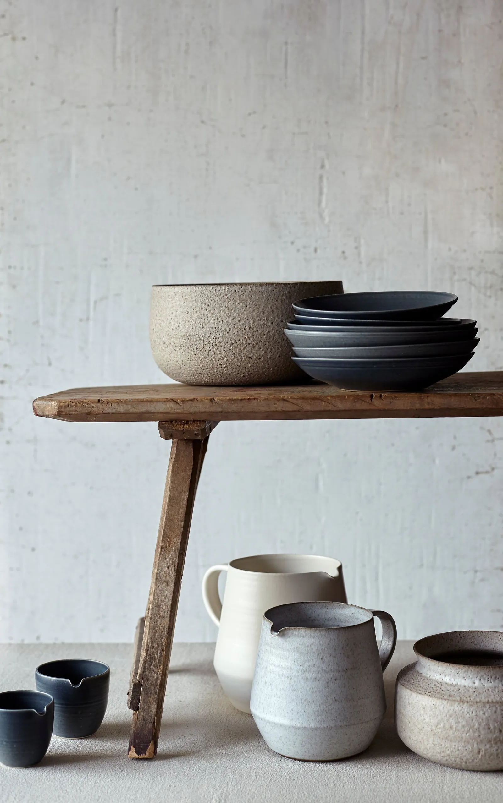 rustic wood bench holding a speckled bowl and stacked navy plates, with cream and grey pitchers and small navy cups on the floor below