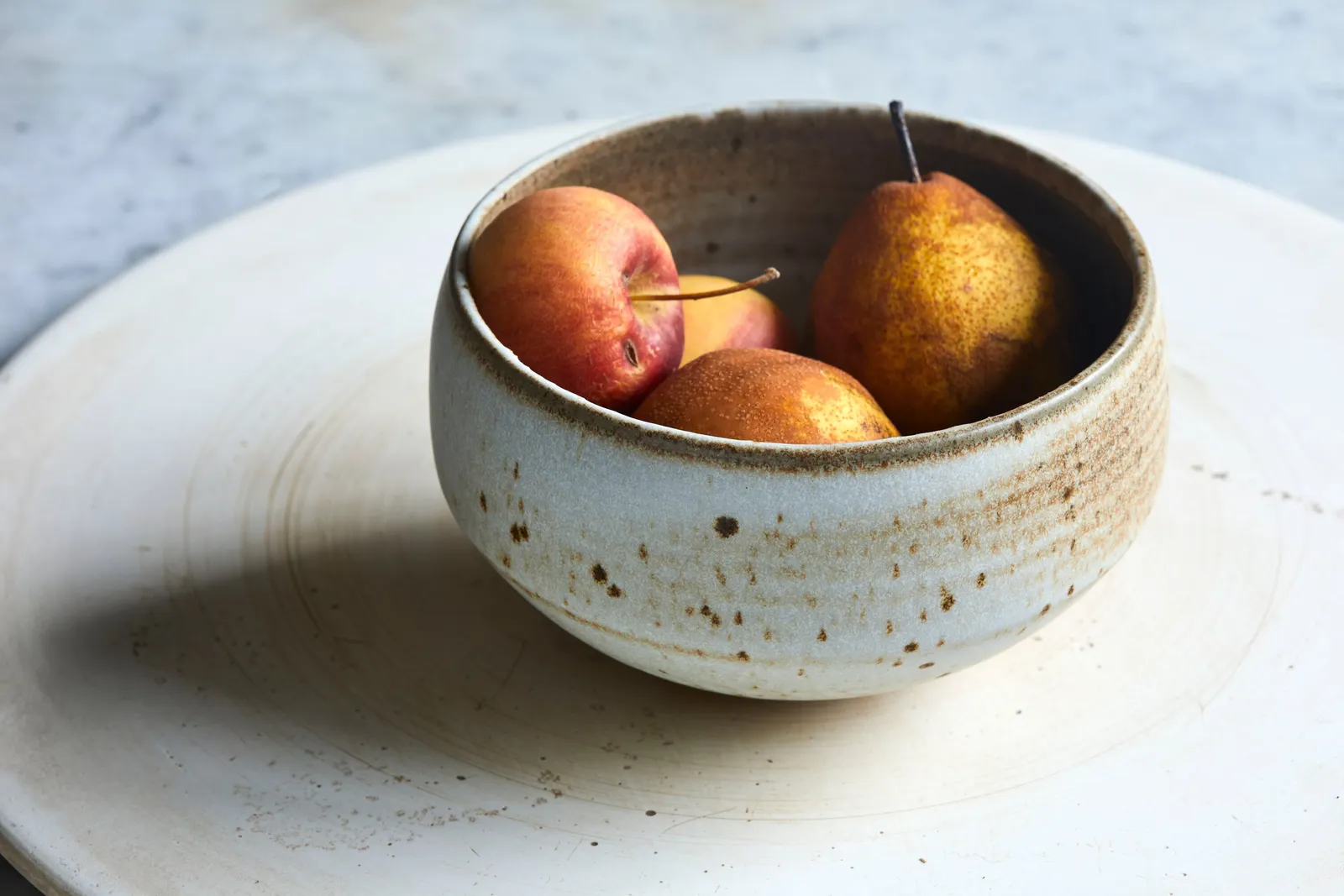 glazed speckled bowl holding two red-yellow apples and a pear on a large cream ceramic platter over a marble surface