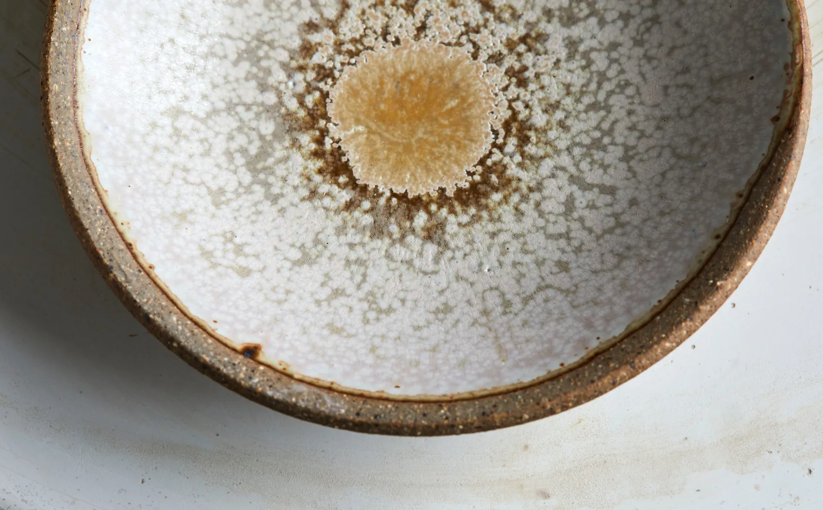 close detail of a bowl with a white crackle glaze, a golden-tan center and a rough brown clay rim on a pale surface