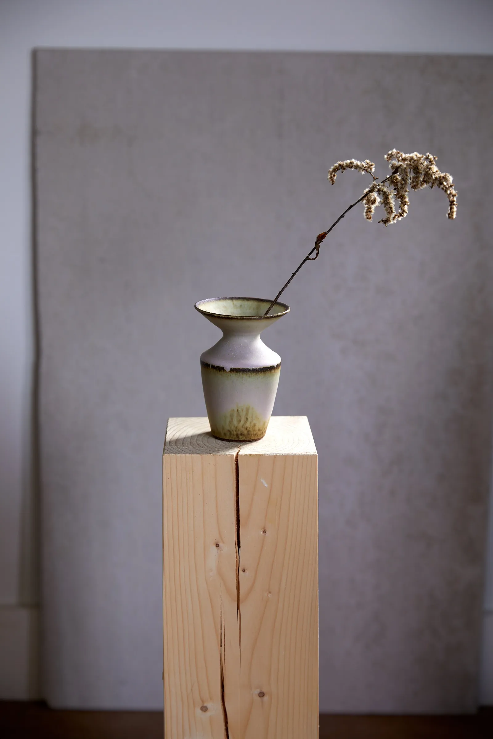 small flared vase with dried goldenrod stem on a raw pine post, against a grey panel and pale wall in soft light
