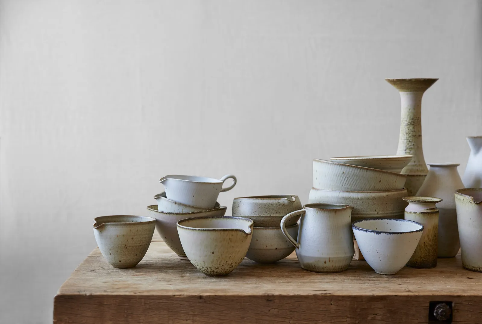 cluster of cream and sand-colored stoneware pitchers, bowls and vases on a rough wooden table against a pale wall