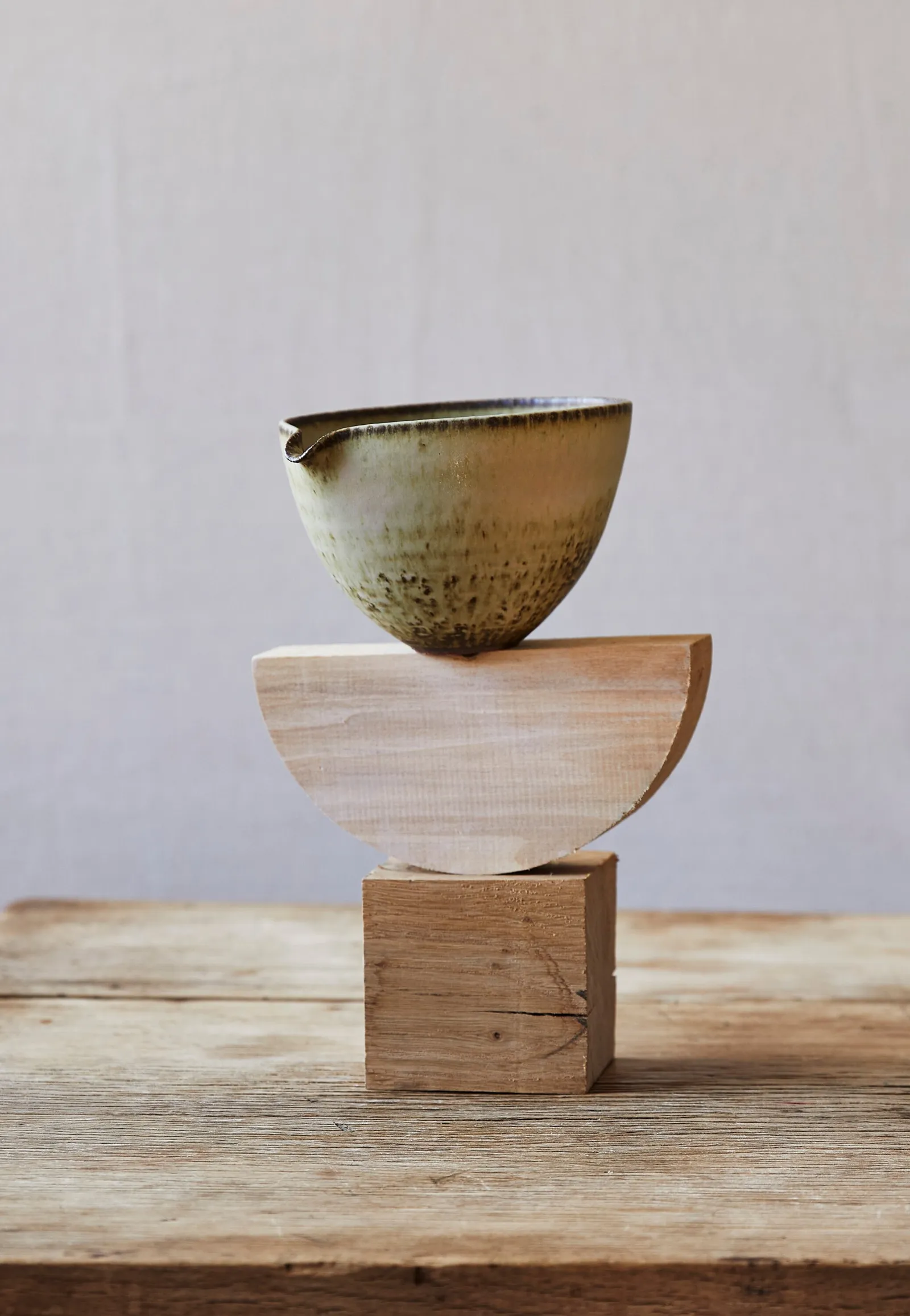 spouted glazed bowl balanced on a half-circle of pale wood stacked on a wood block, on a rough wood table