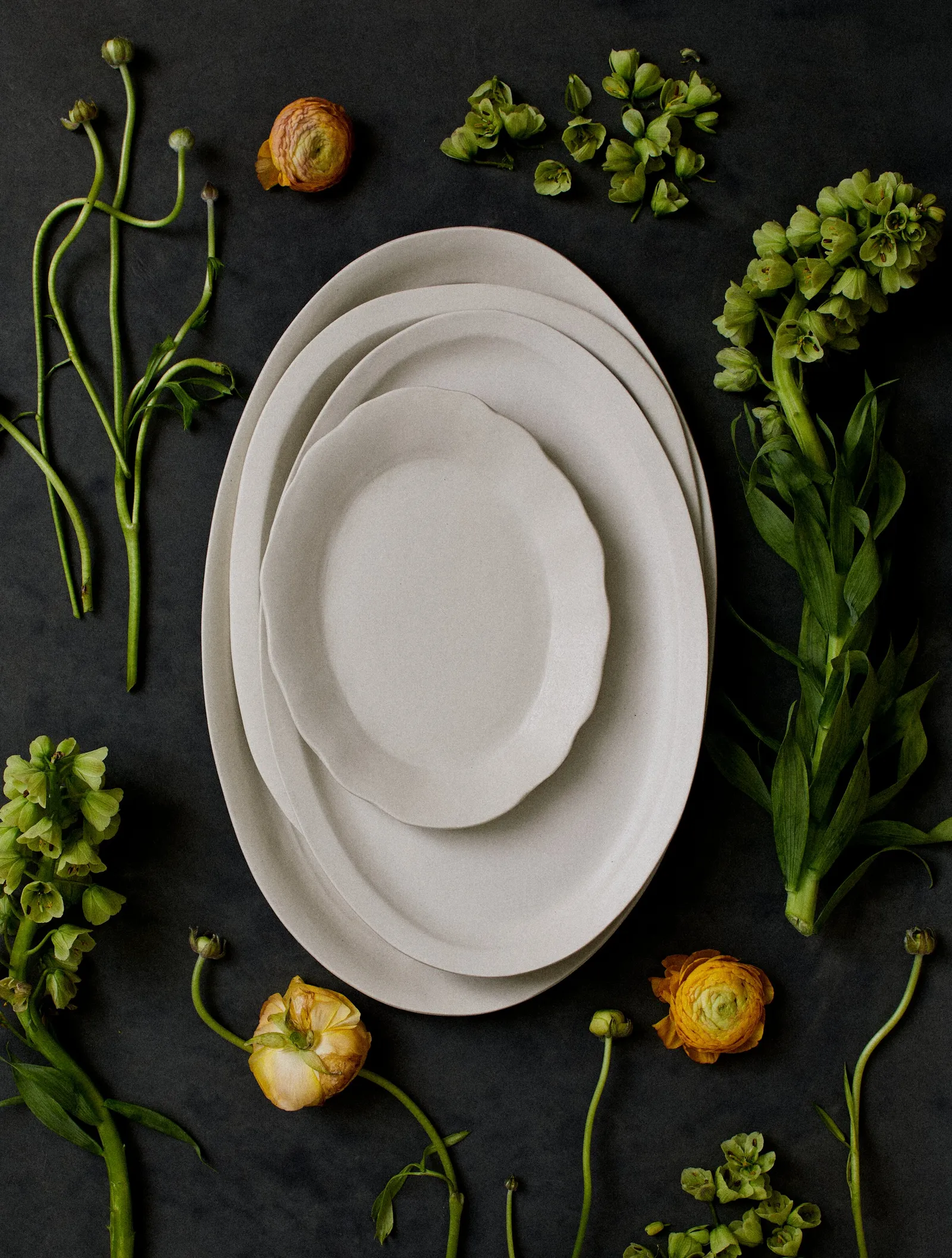 nested cream scalloped platters on a dark slate surface, surrounded by green hellebore stems and orange ranunculus blooms