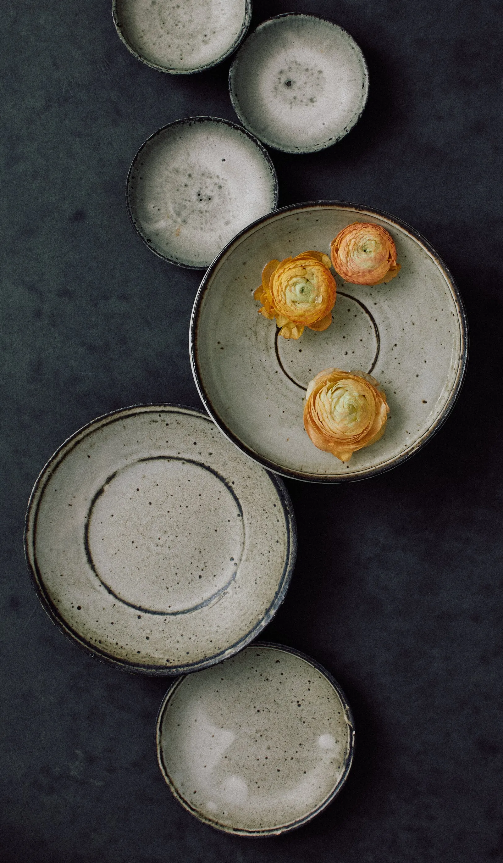 overhead view of speckled grey-green ceramic plates and bowls in a column on dark slate, with three orange ranunculus resting in one bowl
