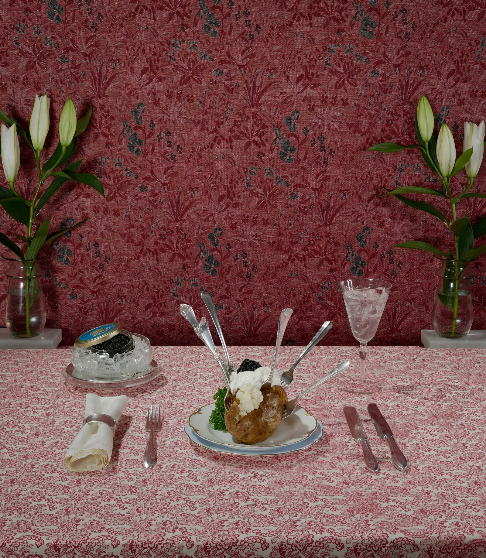 baked potato topped with cream and black caviar, upright silverware around it, with white lilies and a caviar jar on a pink floral tablecloth and wallpaper