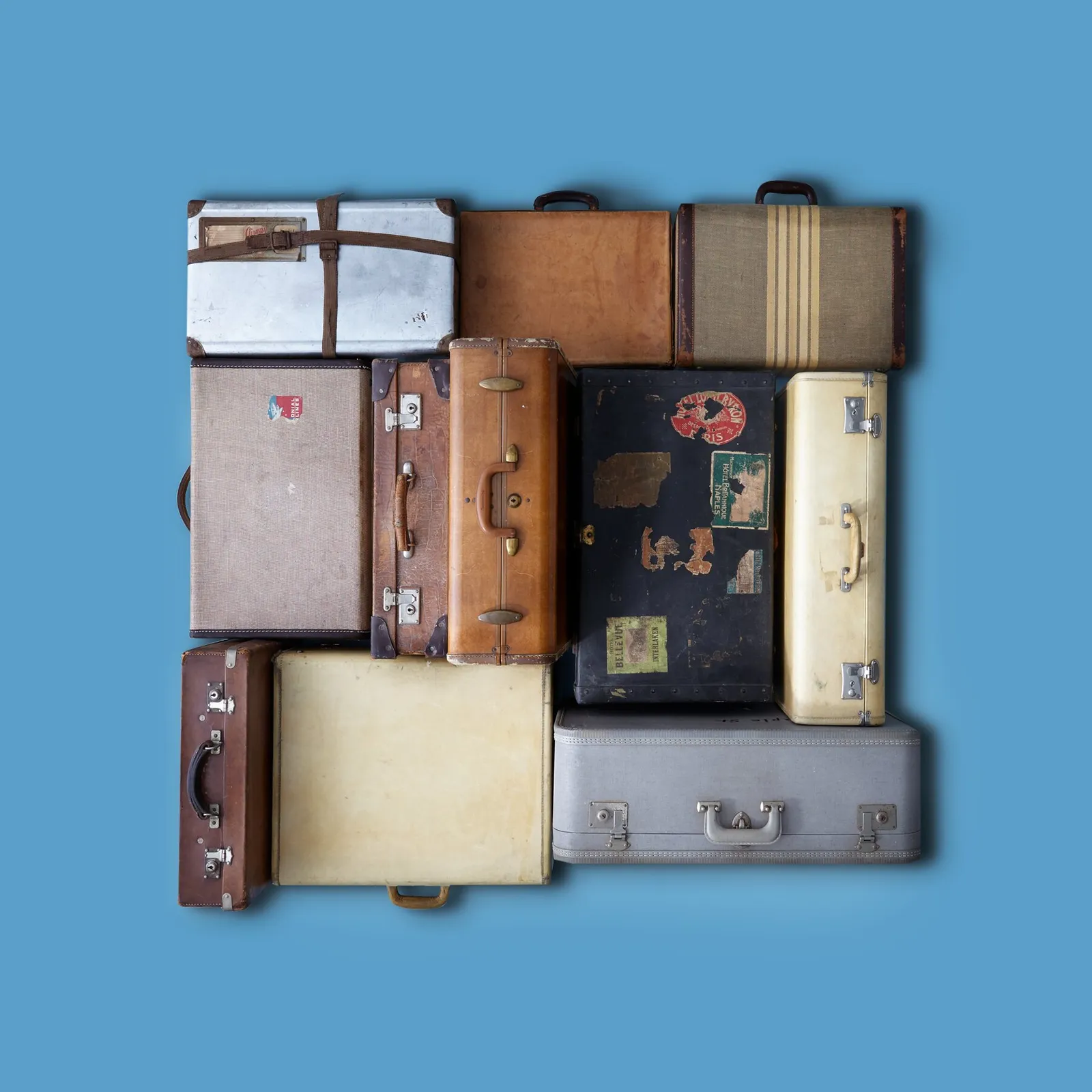 vintage suitcases in tan, cream, black and grey, some with travel stickers, fitted together as a square on a blue background