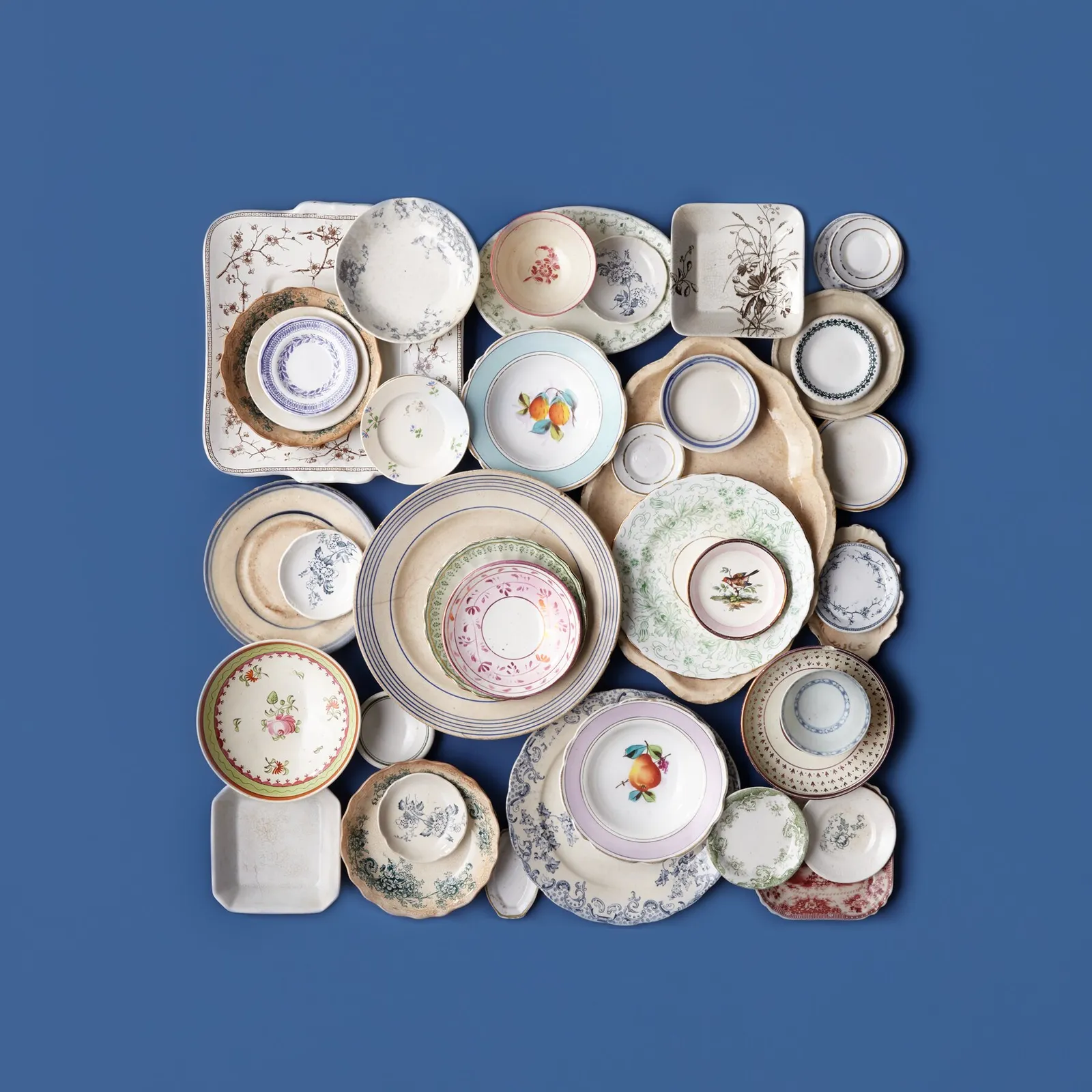 antique china plates and dishes with floral, fruit and bird patterns fitted together in a square on a deep blue background