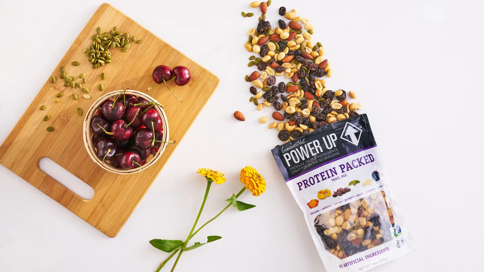 Protein Packed trail mix spilling from its bag beside a bowl of cherries on a cutting board, with pumpkin seeds and two yellow flowers on white