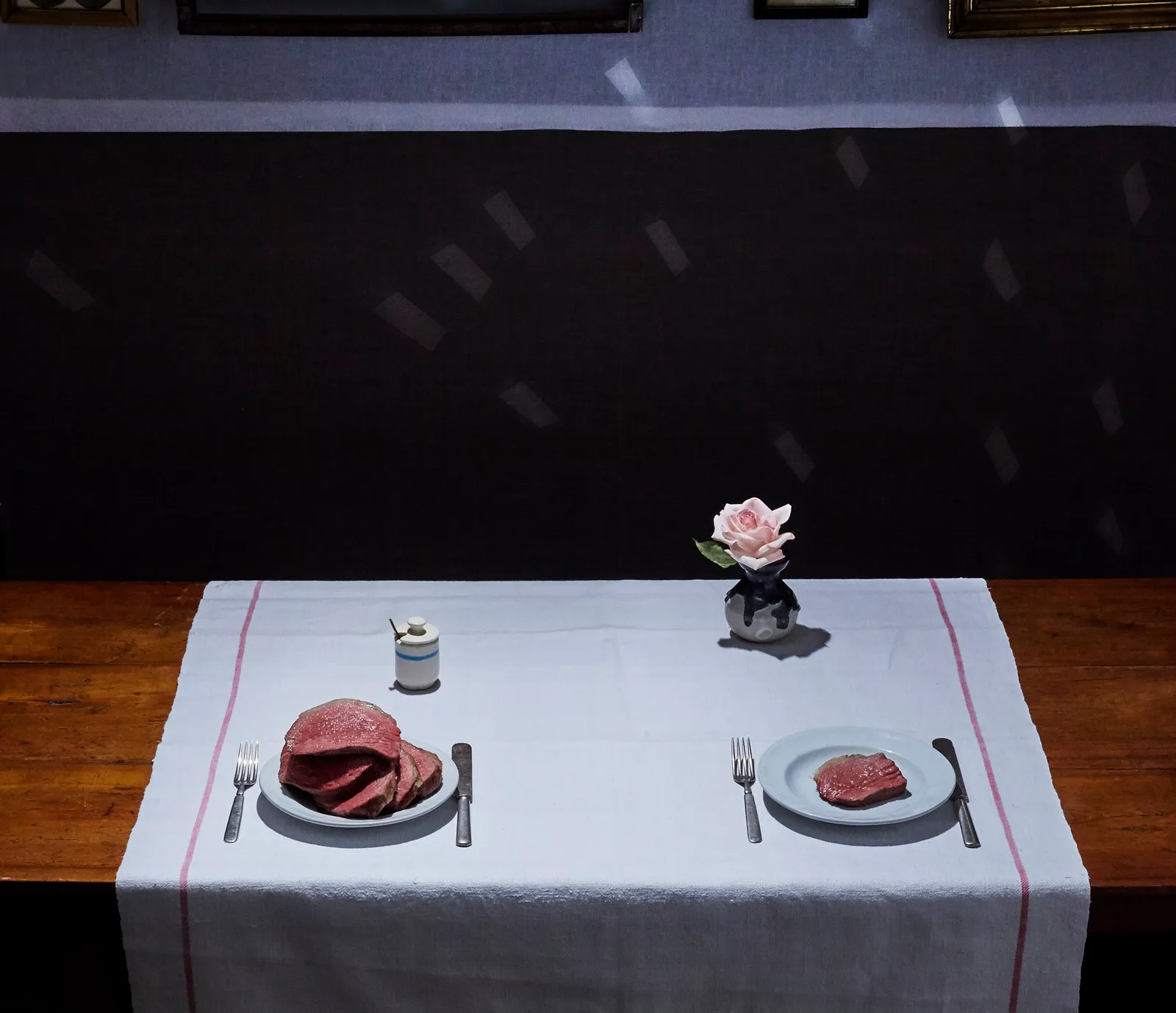 two plates of sliced roast beef, one piled high and one small, with a pink rose in a vase on a white runner over a dark wood table