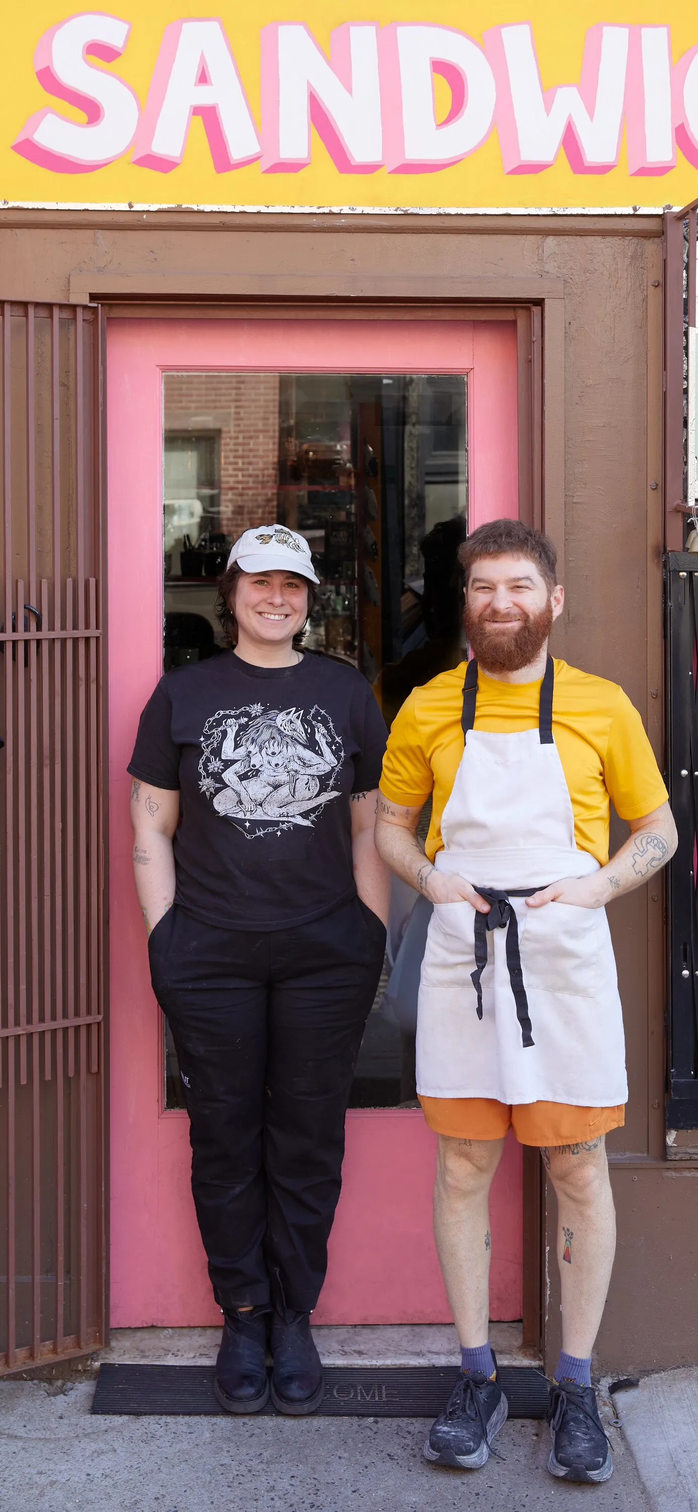 vertical portrait of a smiling woman in a white cap and black t-shirt beside a bearded man in a yellow shirt and apron, in front of a pink door under a yellow sign