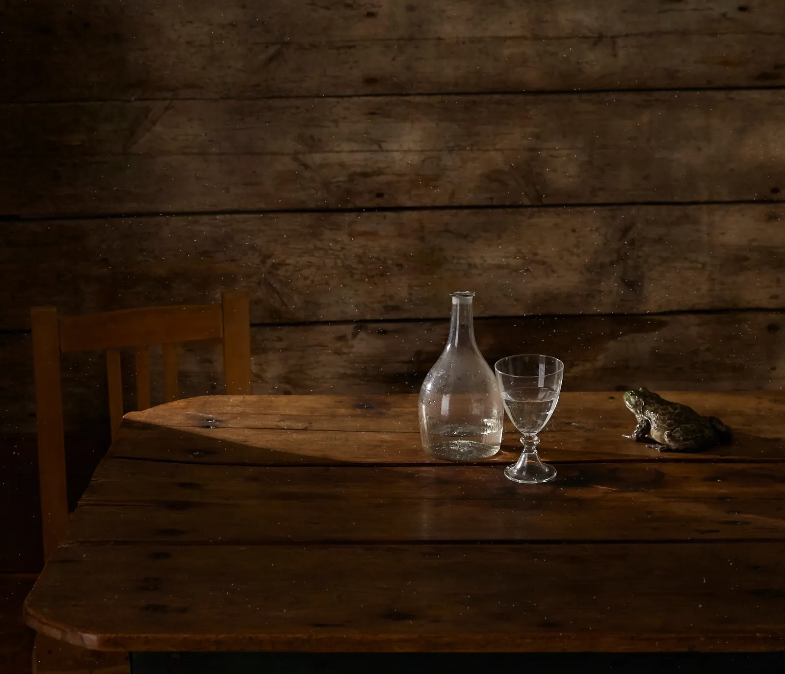 a glass carafe, a goblet of water and a small frog on a rough wood table before a weathered plank wall, with a wooden chair at left