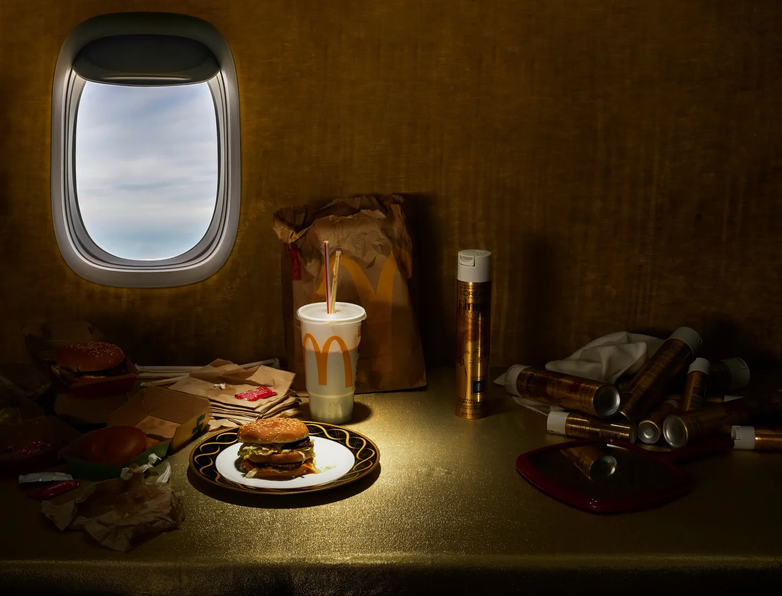 a burger, fries cup and paper bag of fast food lit on a gold-draped table beside hairspray cans, under an airplane window showing clouds