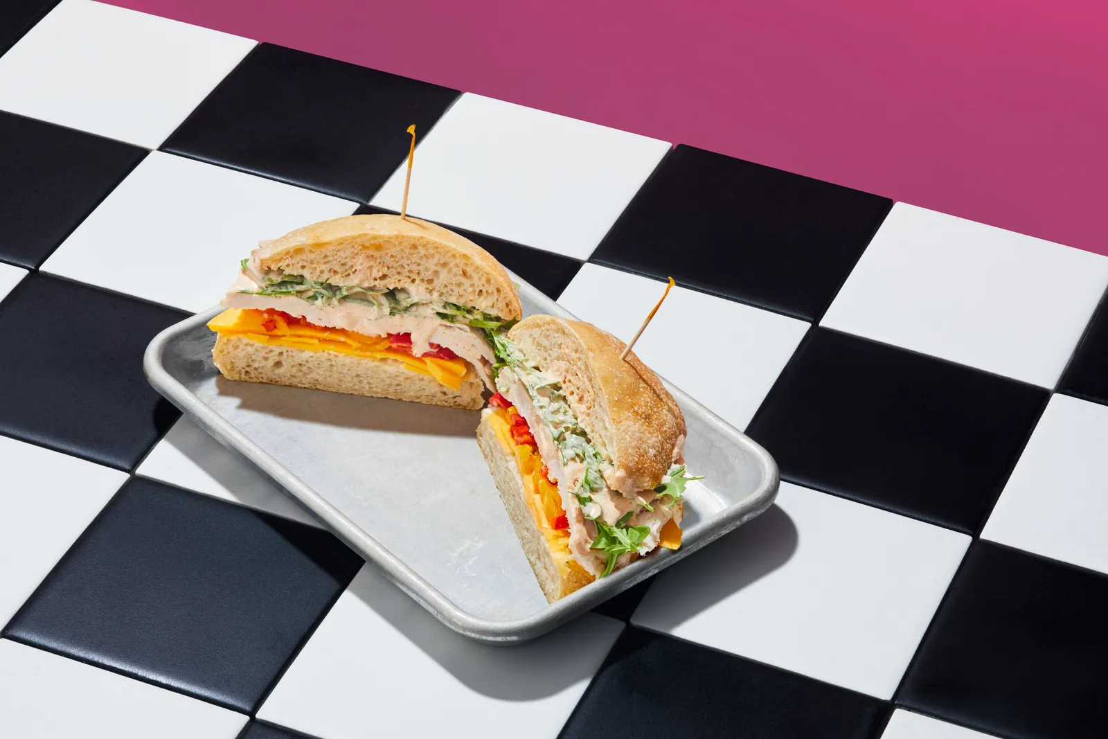 a turkey and cheddar sandwich cut in half with toothpicks on a metal tray, on a black and white checkered floor before a magenta wall