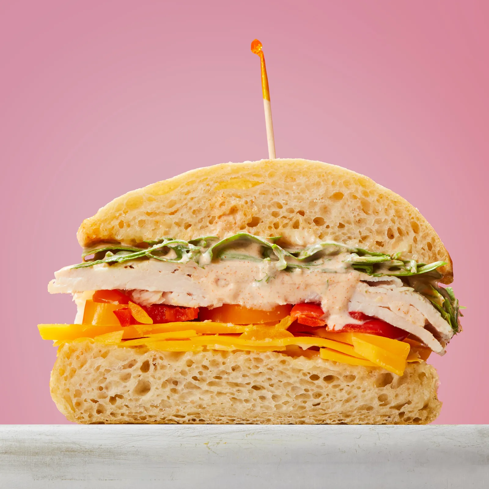 cross-section of a turkey sandwich on ciabatta with cheddar, red pepper, greens and a creamy sauce, against a pink background