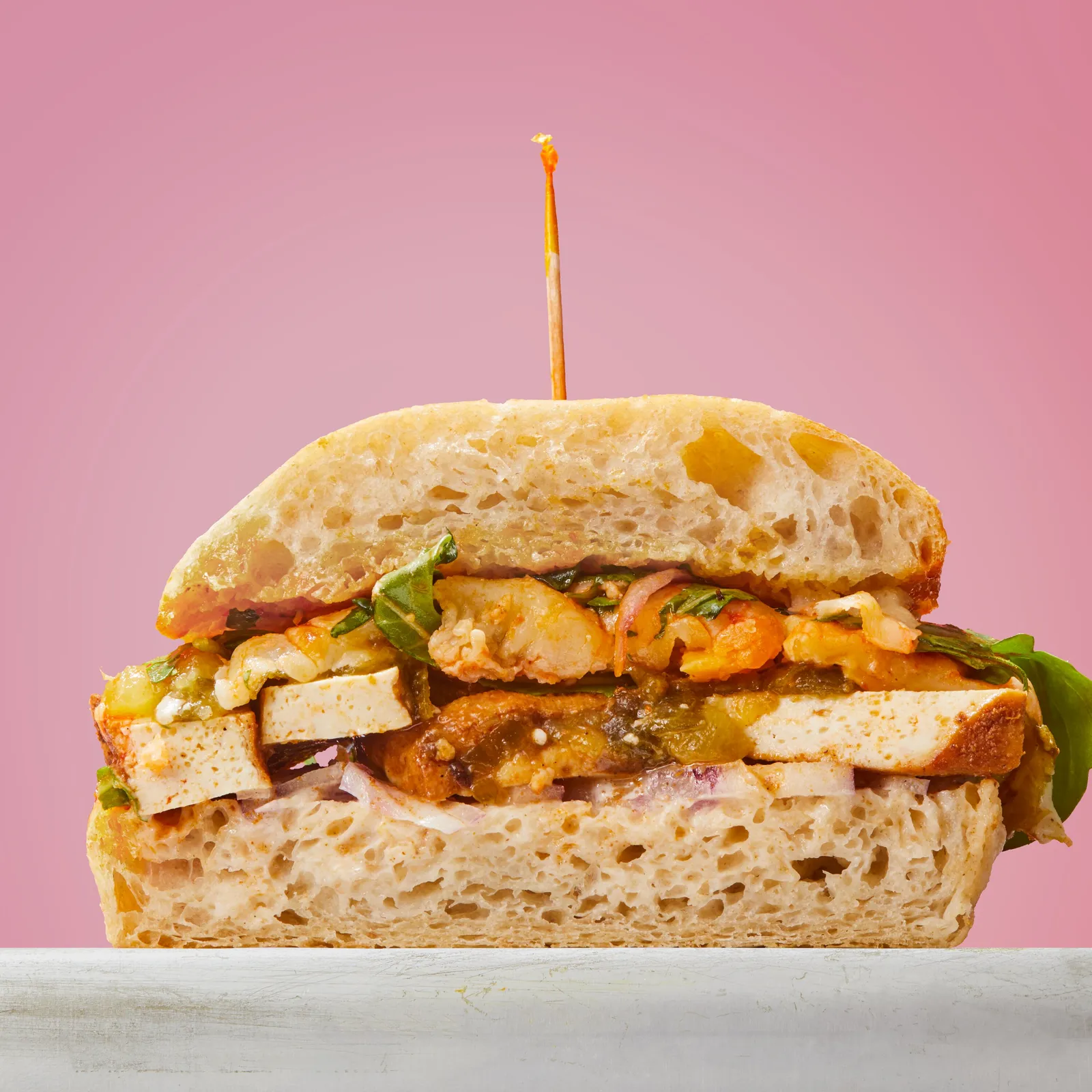 cross-section of a ciabatta sandwich with tofu slices, roasted orange and green vegetables, red onion and arugula, against pink