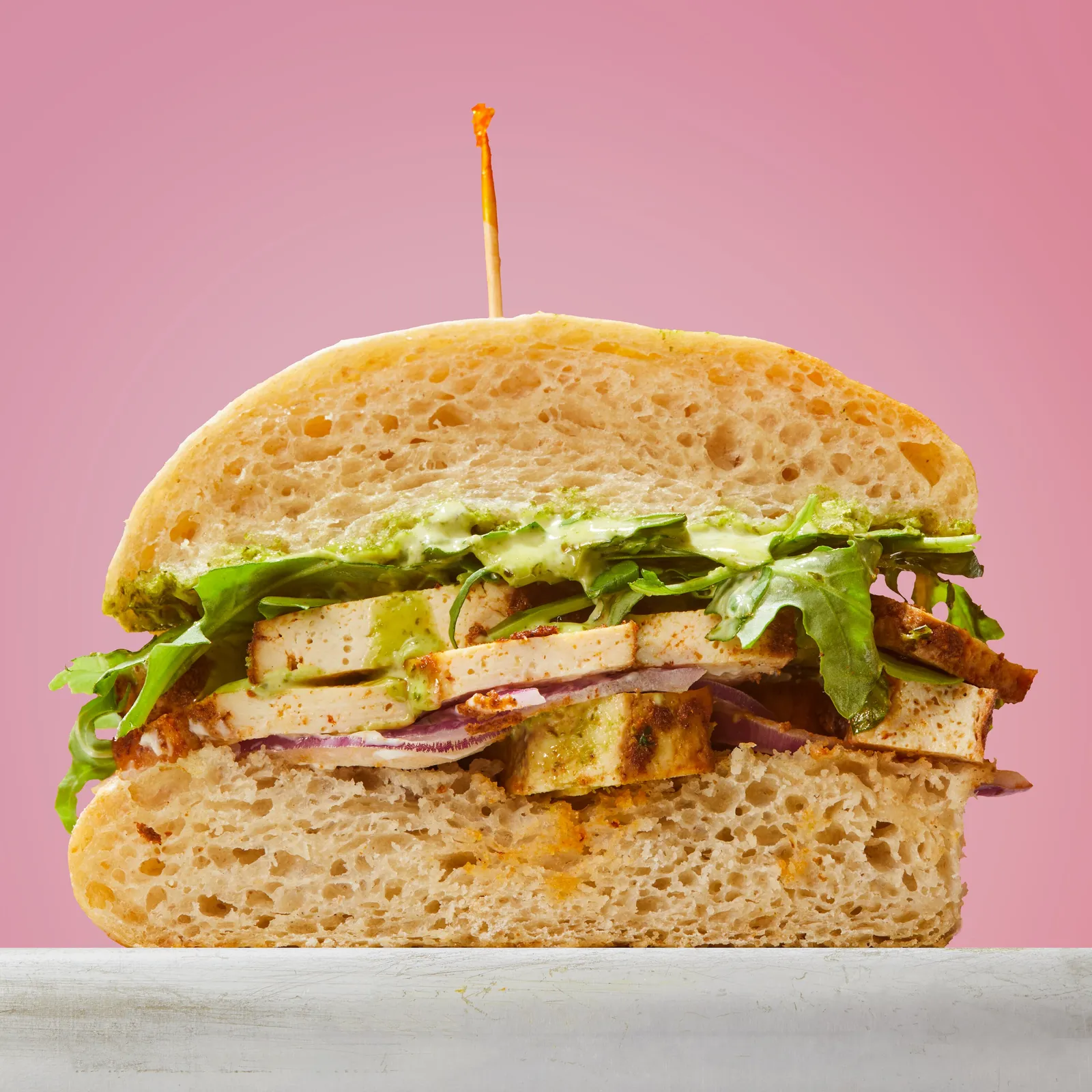 cross-section of a sandwich with seared tofu, green herb sauce, arugula and red onion on ciabatta, against pink