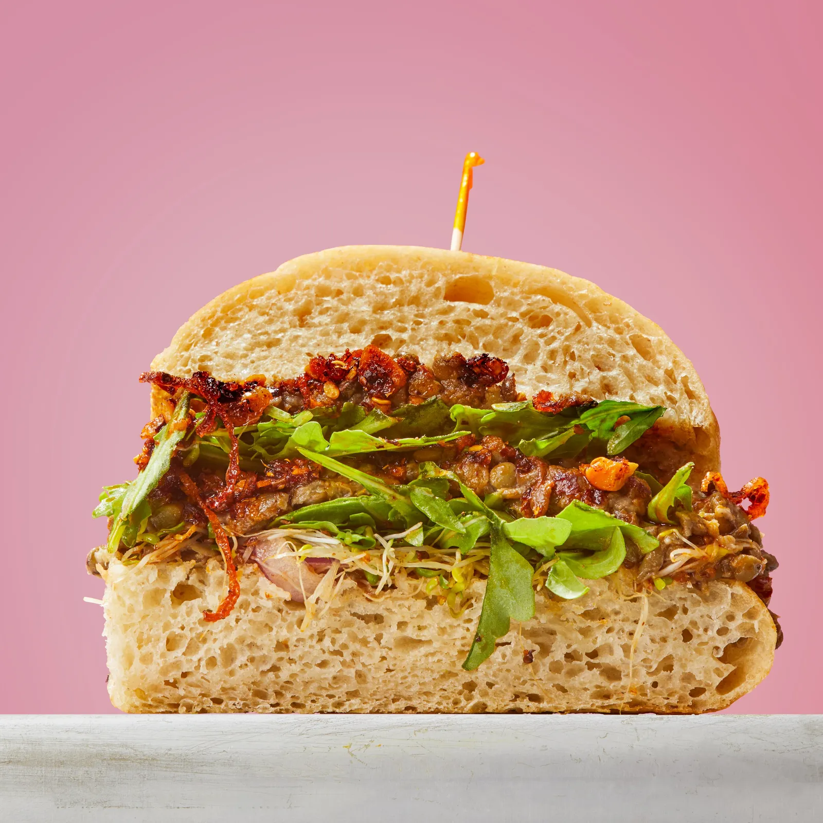 cross-section of a sandwich piled with lentils, arugula, sprouts and crispy chili on ciabatta, against pink