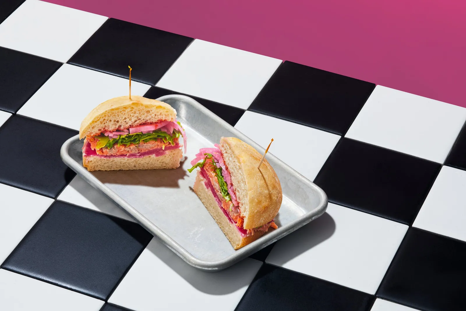 a sandwich with pink pickled onions and greens cut in half with toothpicks on a metal tray, on a checkered floor