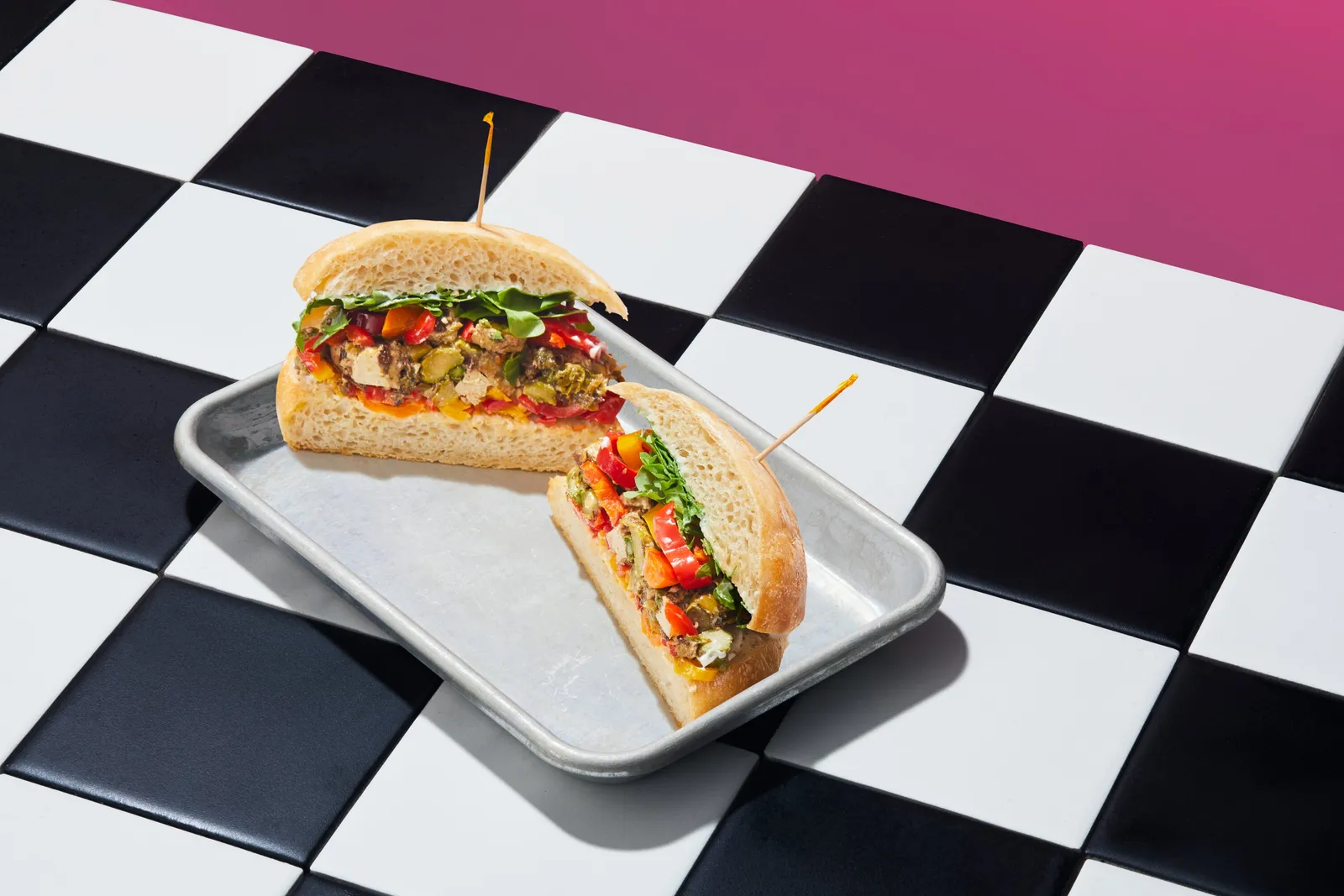 a sandwich with roasted peppers and green vegetables cut in half with toothpicks on a metal tray, on a checkered floor