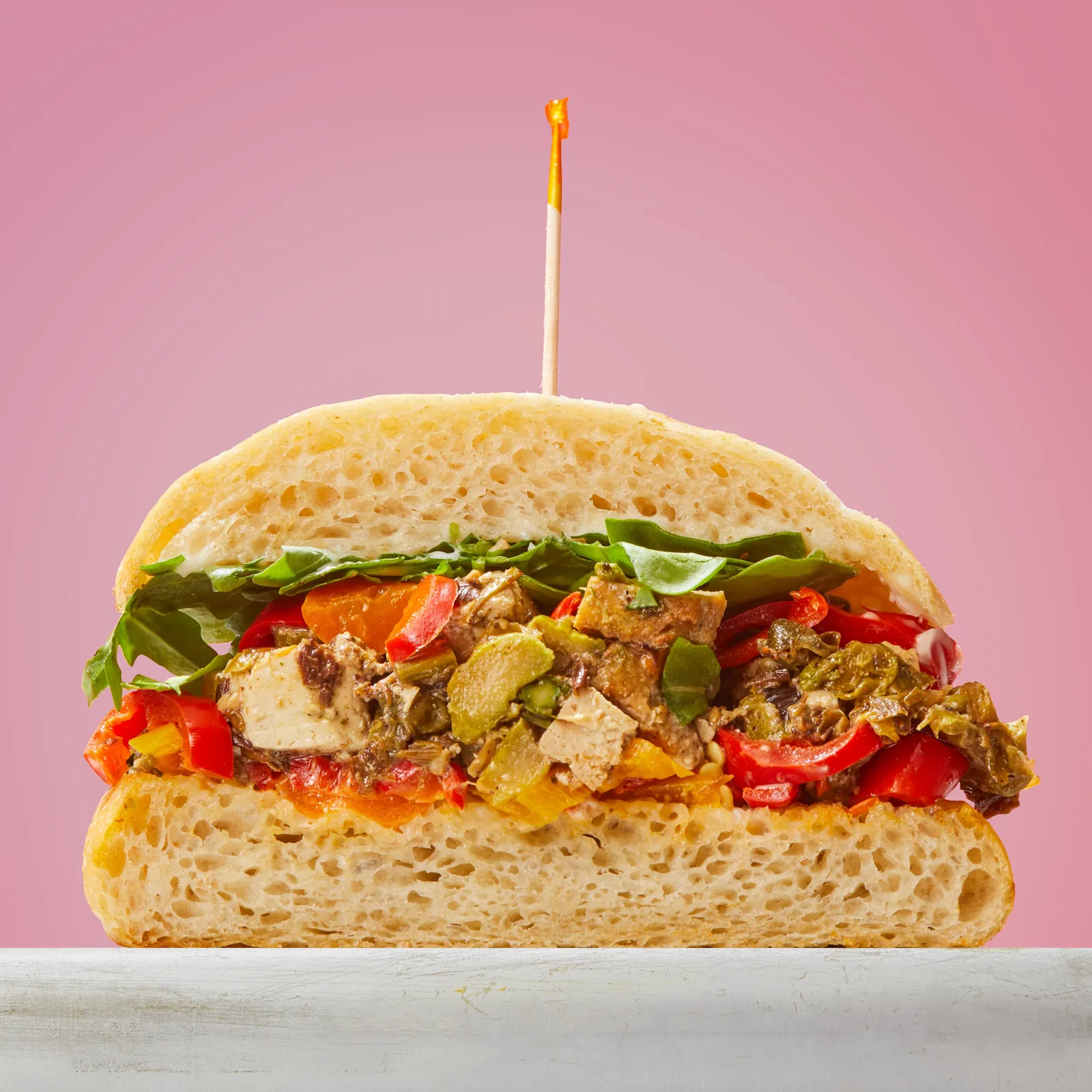 cross-section of a sandwich with roasted red peppers, tofu, greens and olives on ciabatta, against pink