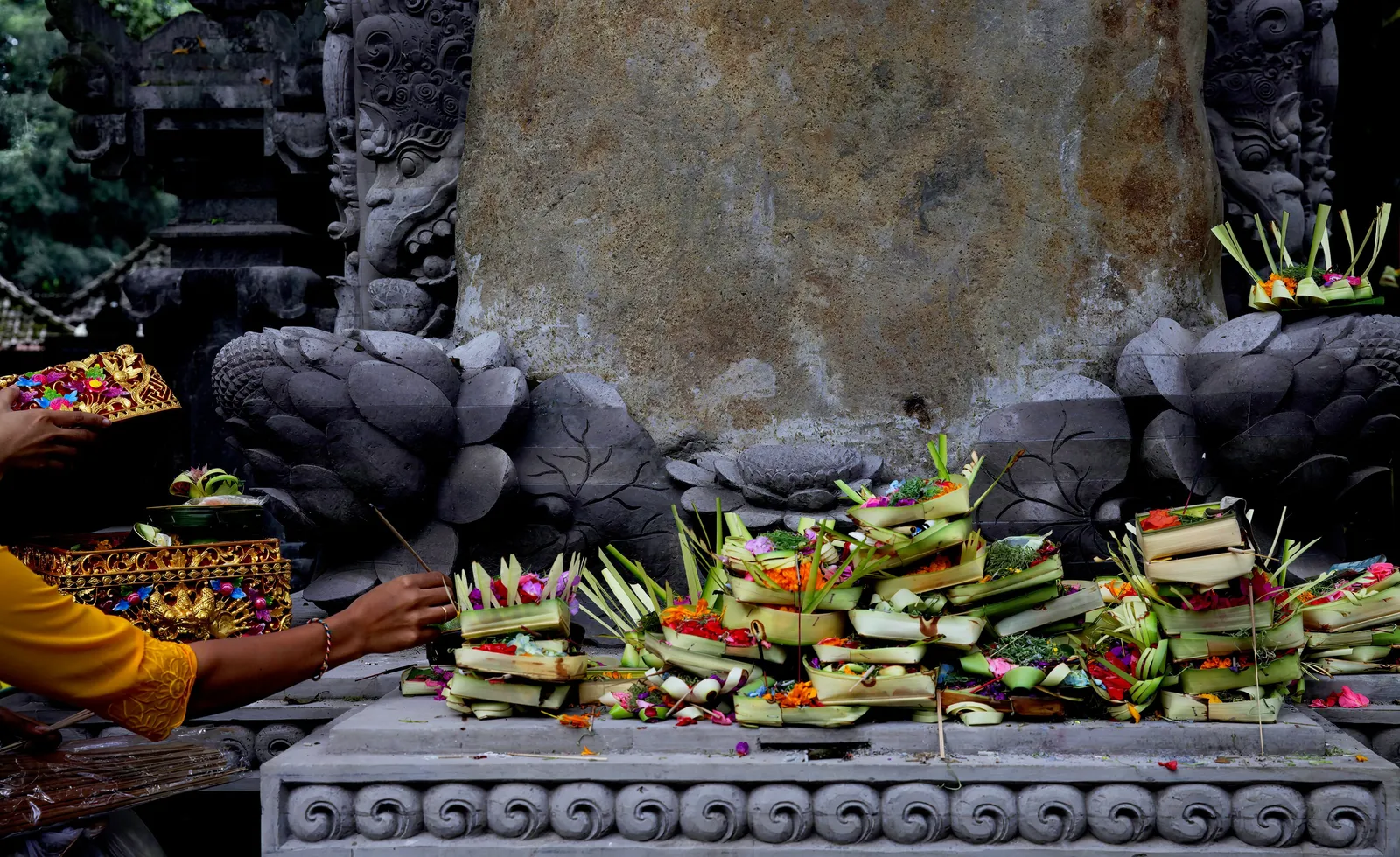 colorful flower and leaf offerings piled on a carved stone ledge, with a woman in a yellow sleeve arranging them before a weathered stone relief