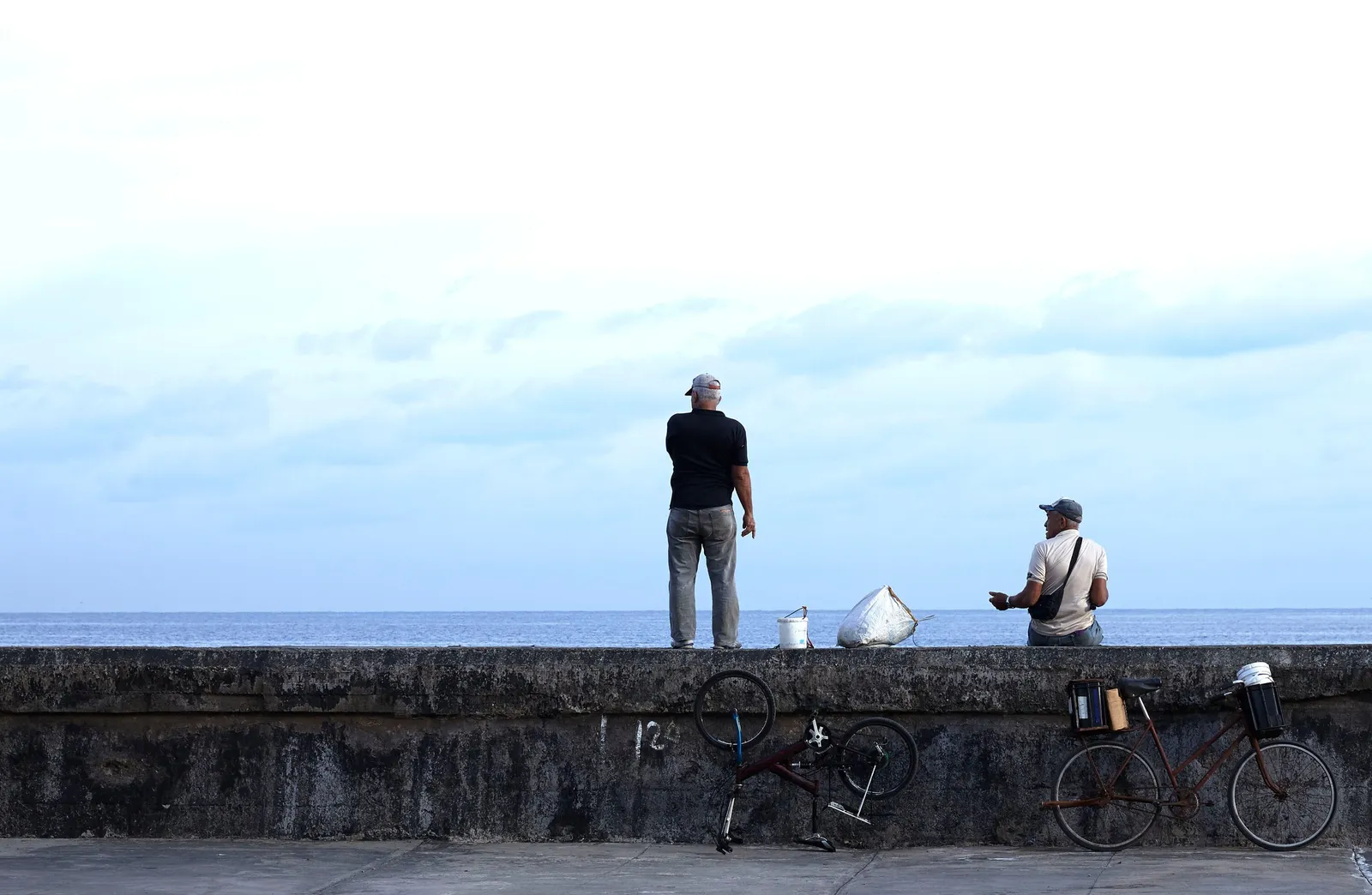 two men on a seawall, one standing and one seated, with bicycles leaning against it and the sea and pale blue sky behind