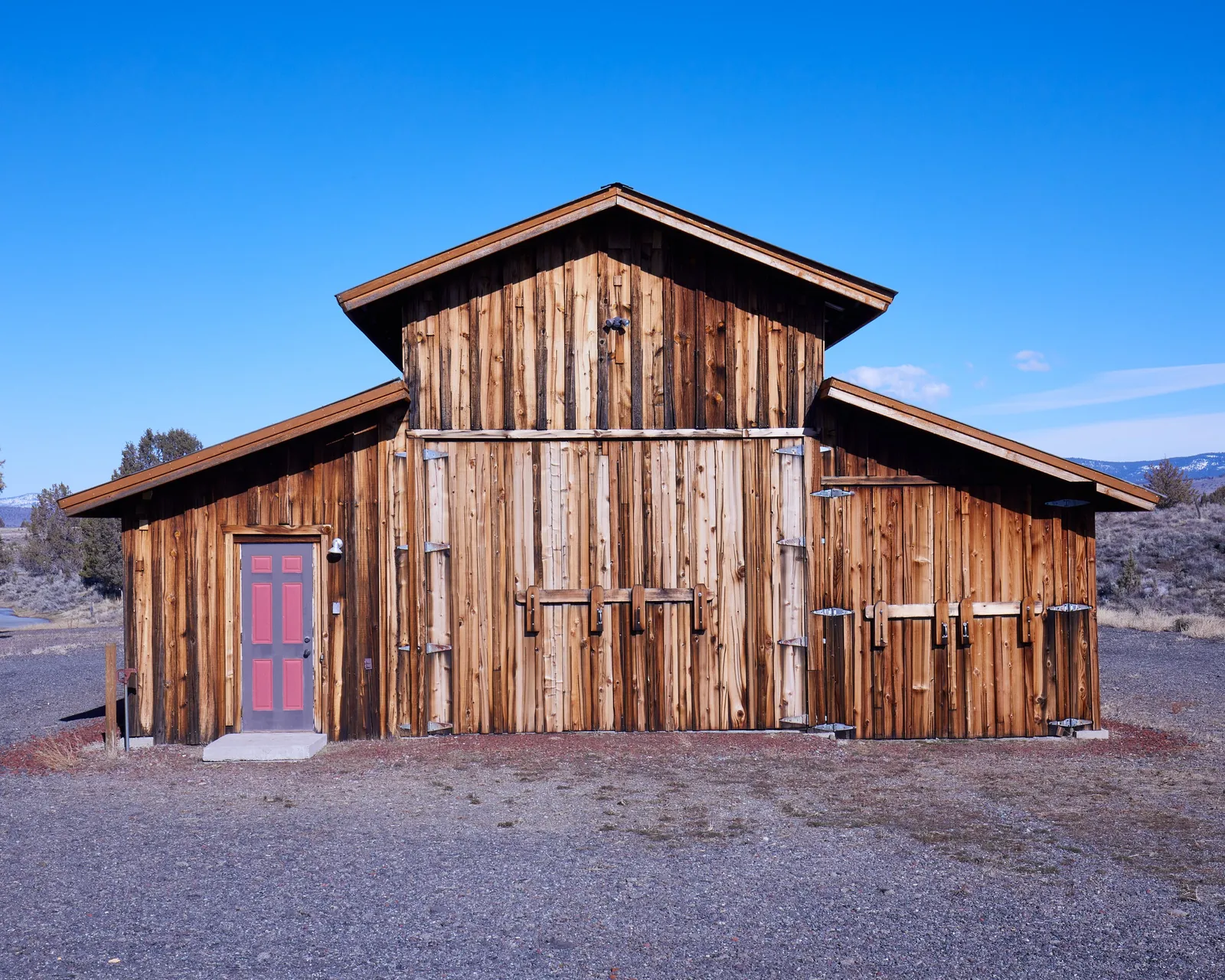 weathered wood barn with a pink door, centered under a bright blue sky with gravel in front and low hills behind