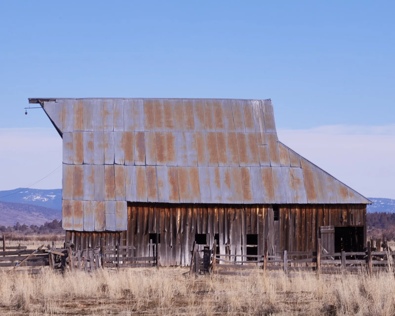 old barn with a rusted corrugated metal roof over gray wood siding, set in dry grass with distant snowy hills