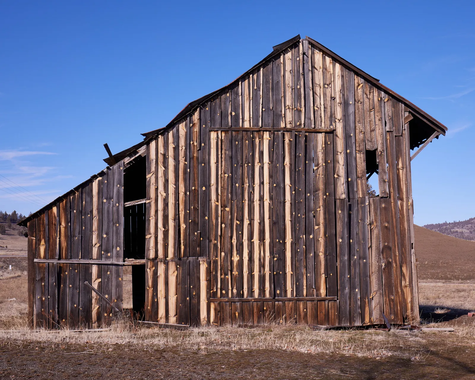 side of a leaning wood barn with weathered vertical boards and open gaps near the roofline, against deep blue sky