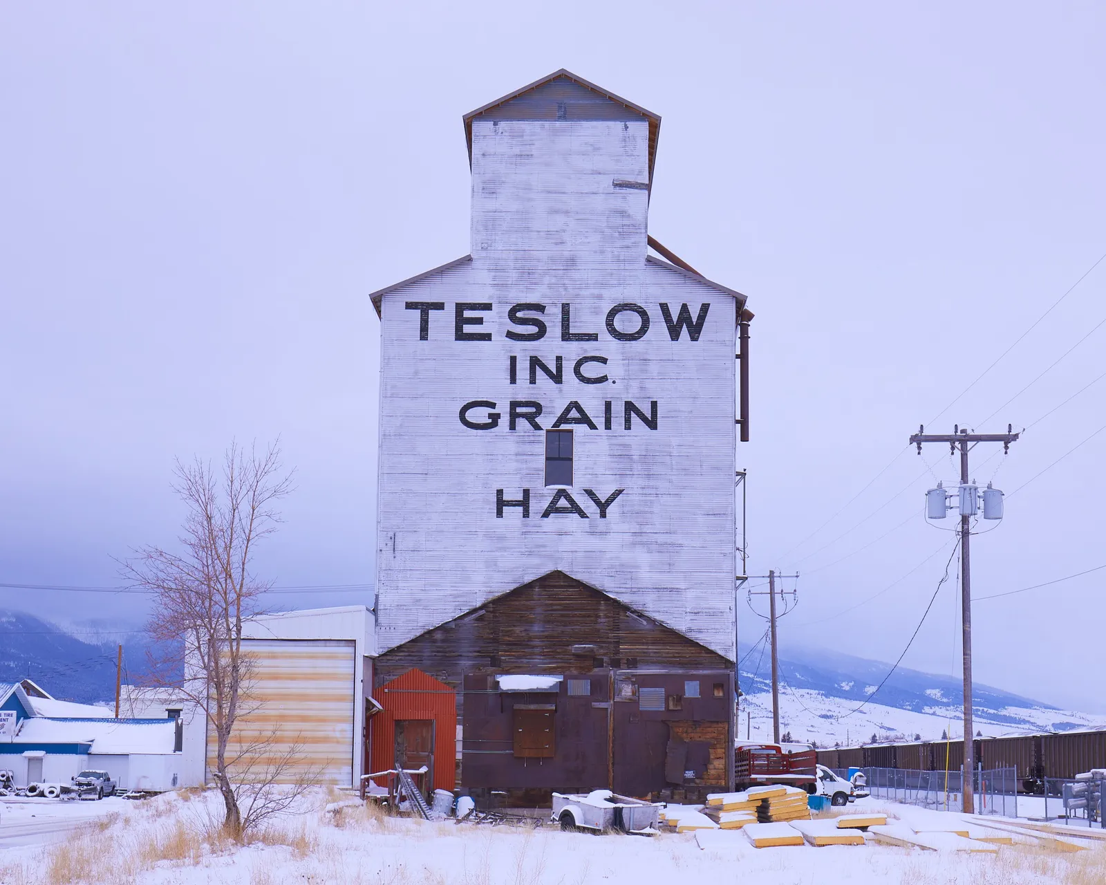 white grain elevator painted with the words Teslow Inc. Grain Hay, standing in snow under a pale overcast sky with power lines