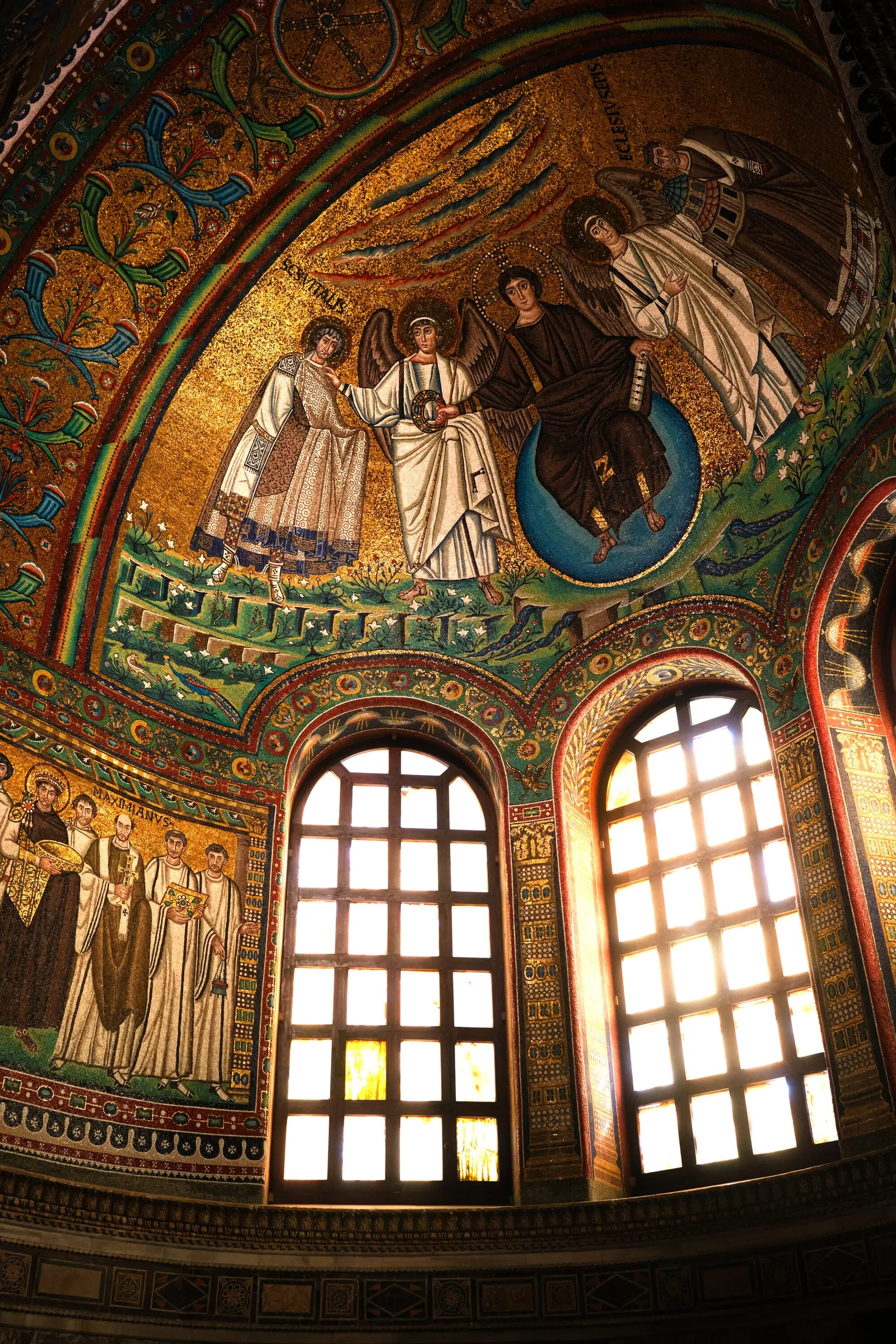 vertical view inside a church apse covered in gold mosaics of robed figures and angels, with two tall lit windows below