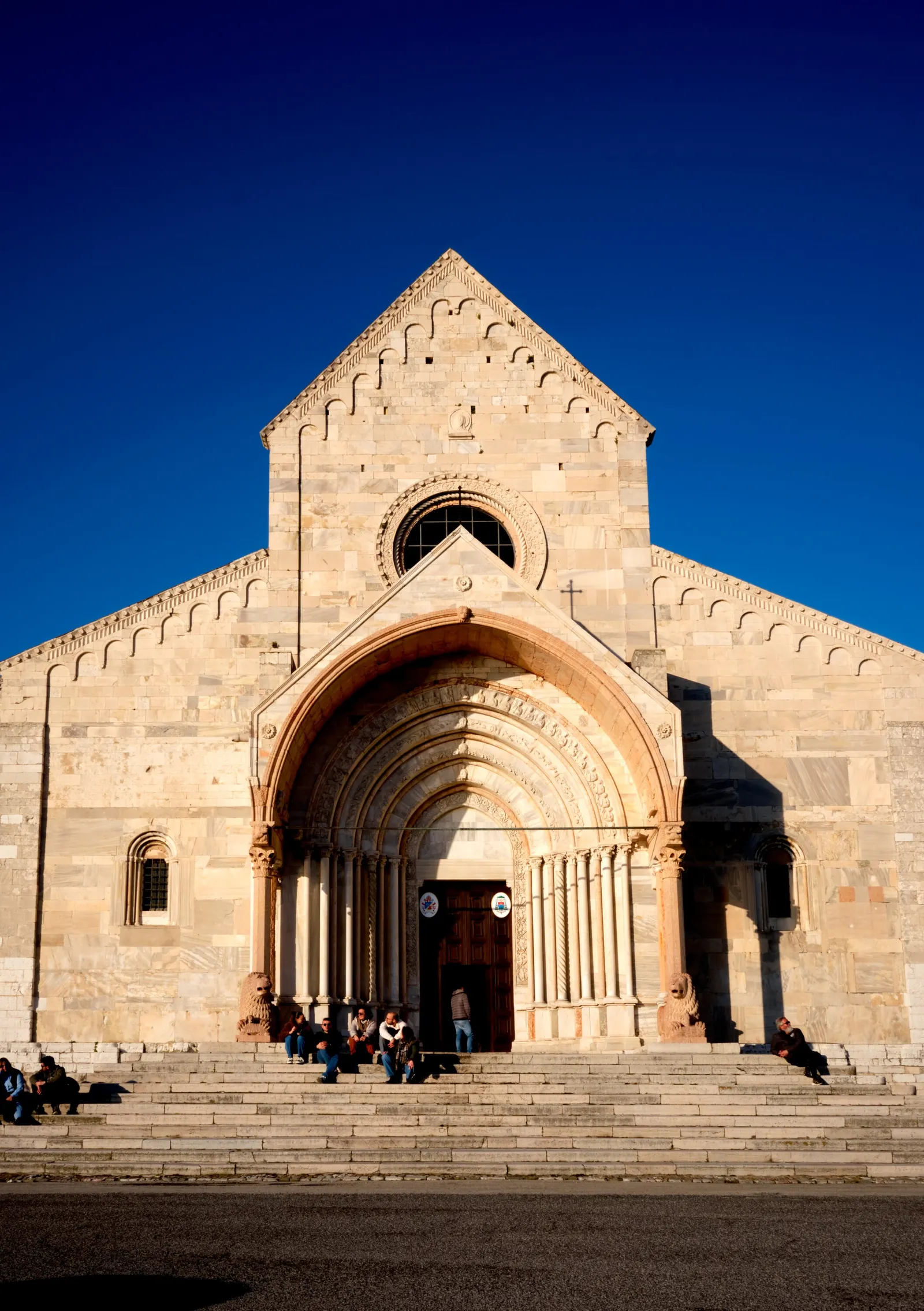 stone church facade with a pointed gable, round window and layered arched doorway, people sitting on the wide steps under a deep blue sky