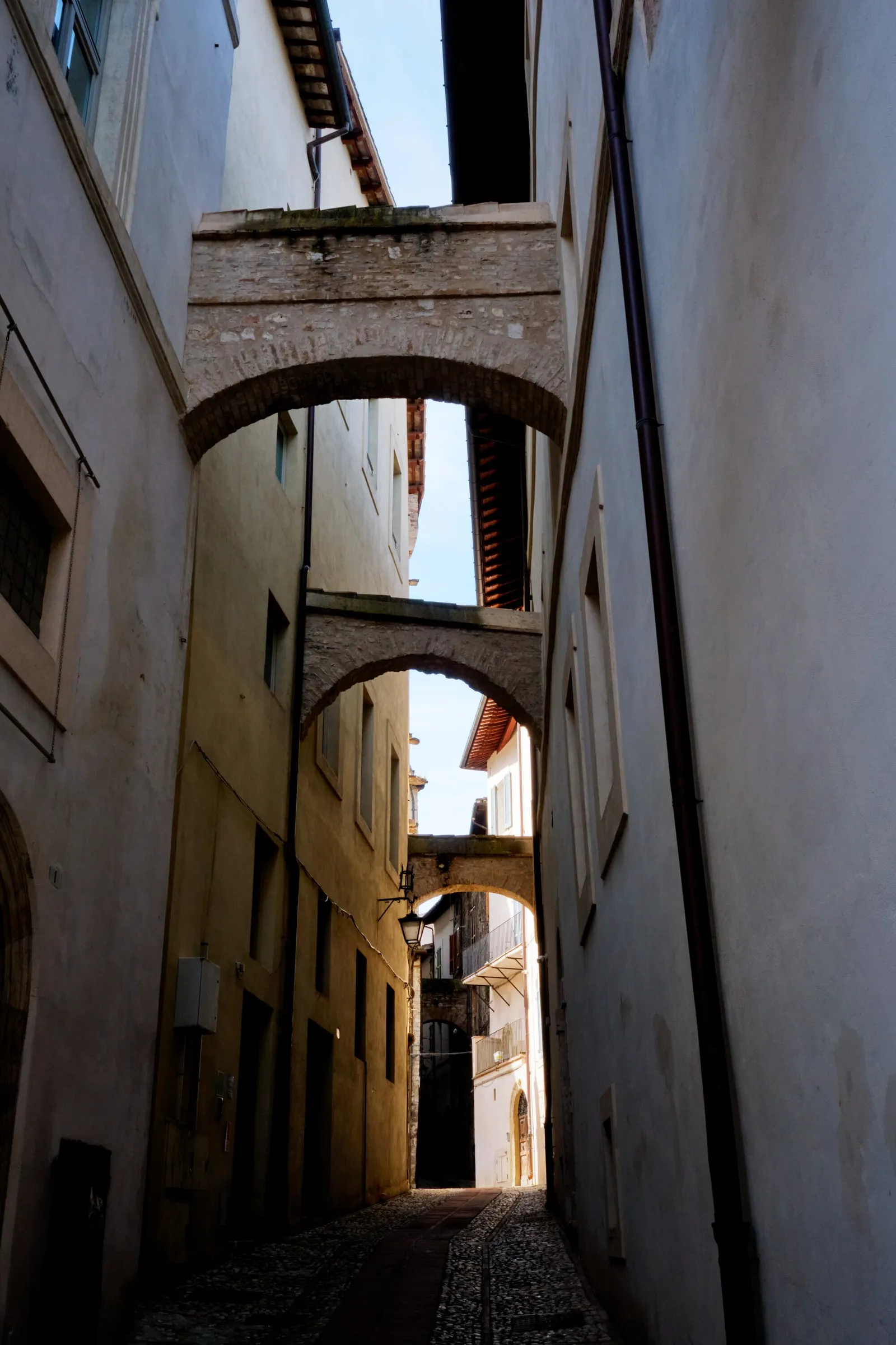 narrow cobbled lane between pale plaster buildings, crossed by two stone arches with a sliver of blue sky above