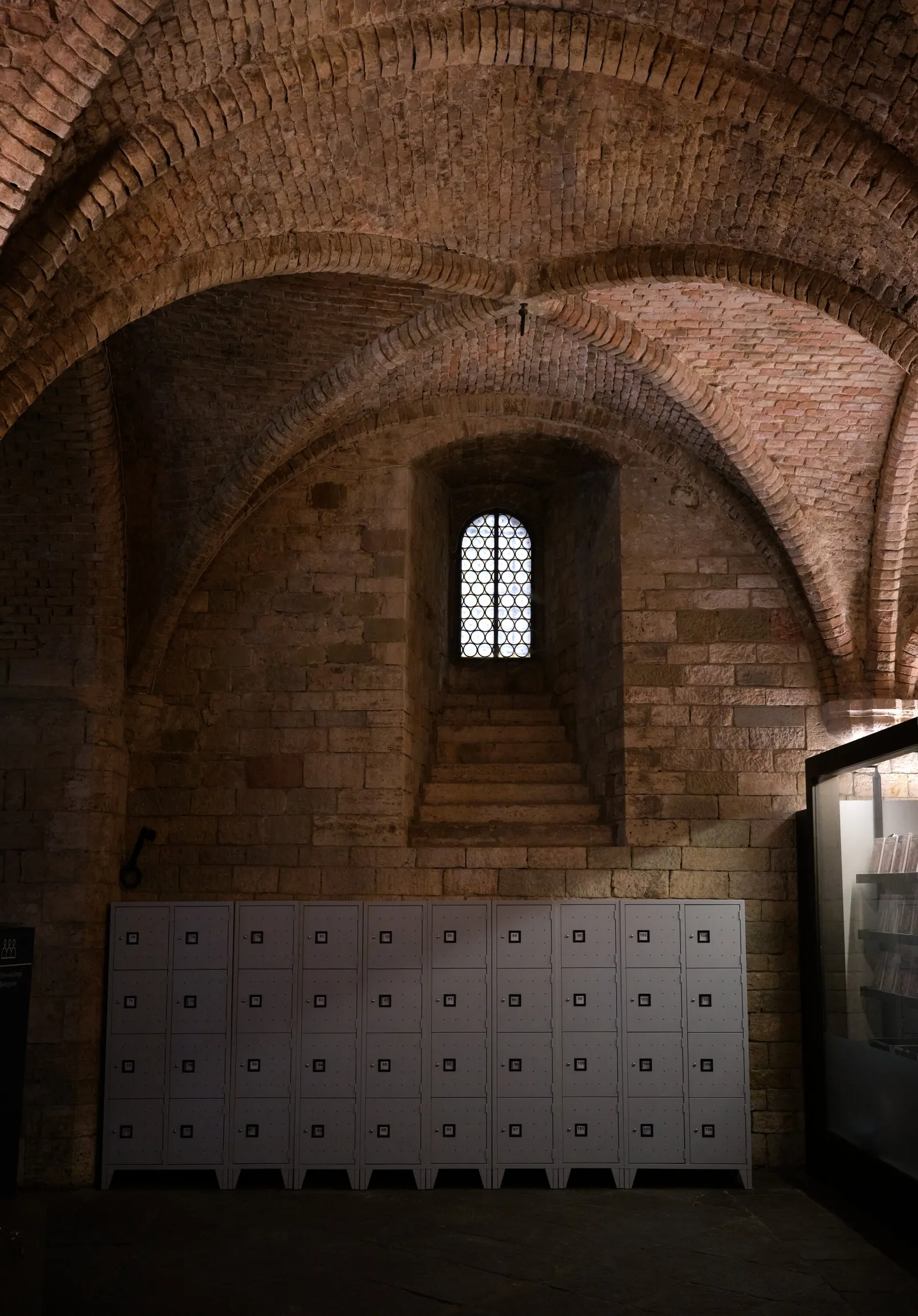 dim brick vaulted hall with a small lattice window at the top of a stair niche and a row of gray lockers along the wall