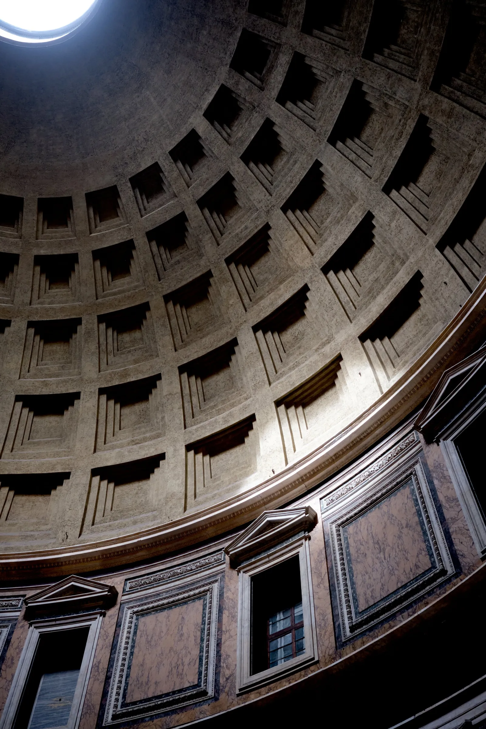 looking up into a coffered dome with a bright oculus at top left and a shaft of light falling across the ceiling and marble wall below