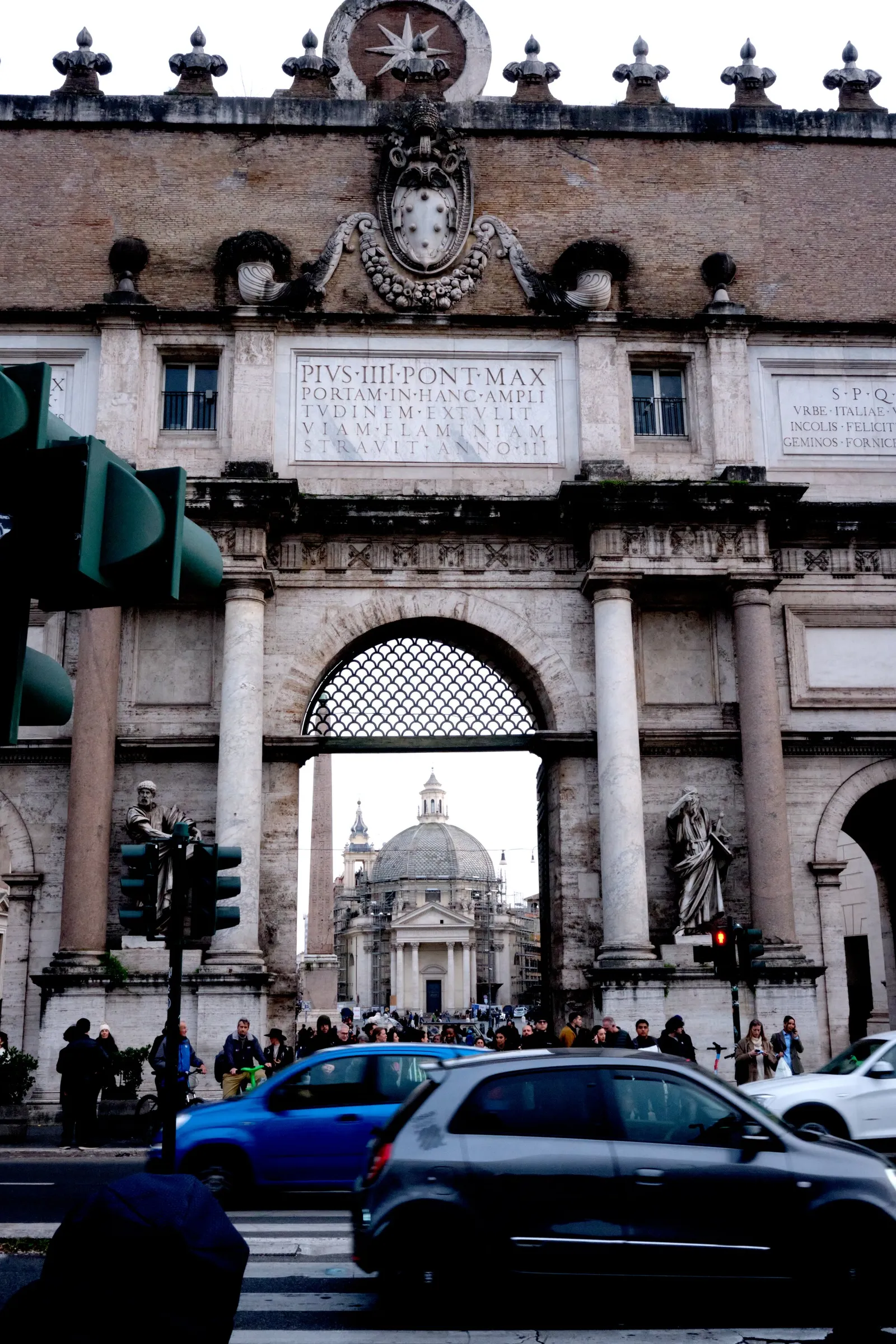 carved stone city gate with an inscribed panel and statues, a domed church seen through the arch, traffic and pedestrians in the foreground