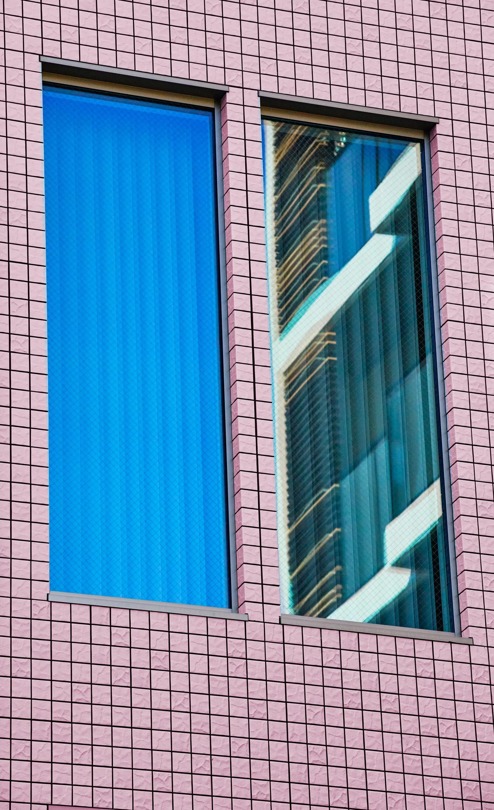 two tall narrow windows in a wall of pink square tiles, one filled with a flat blue curtain and the other reflecting a stepped building in warm light