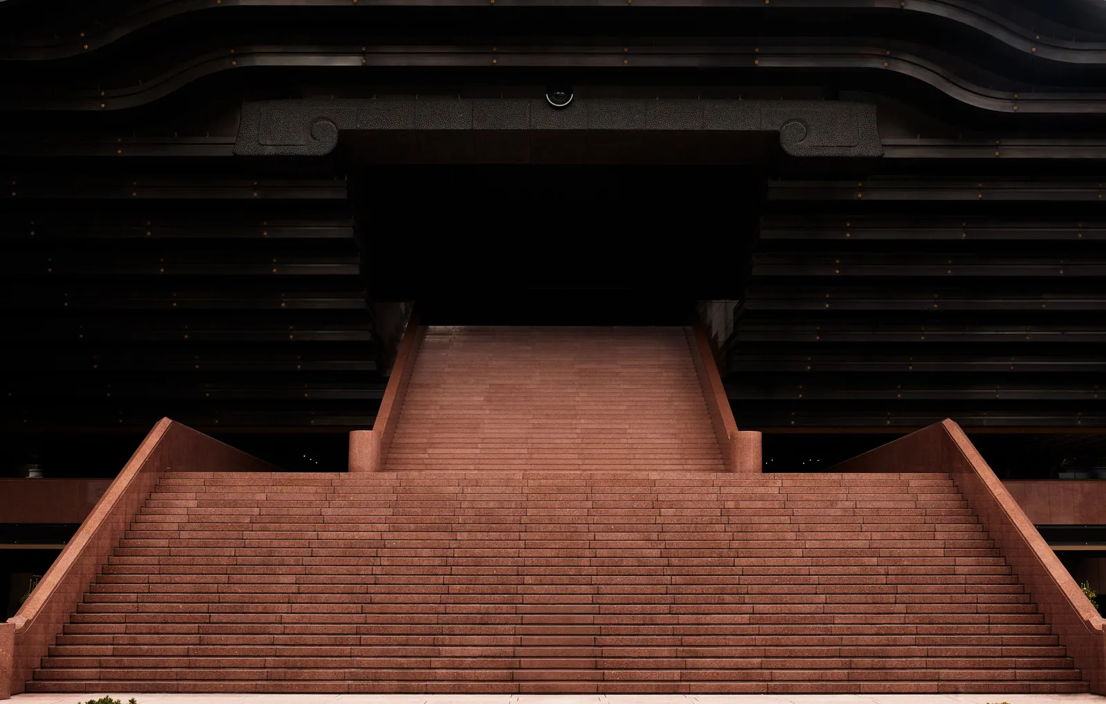 wide red stone staircase rising to a dark recessed entrance beneath a black curved facade, strongly symmetrical