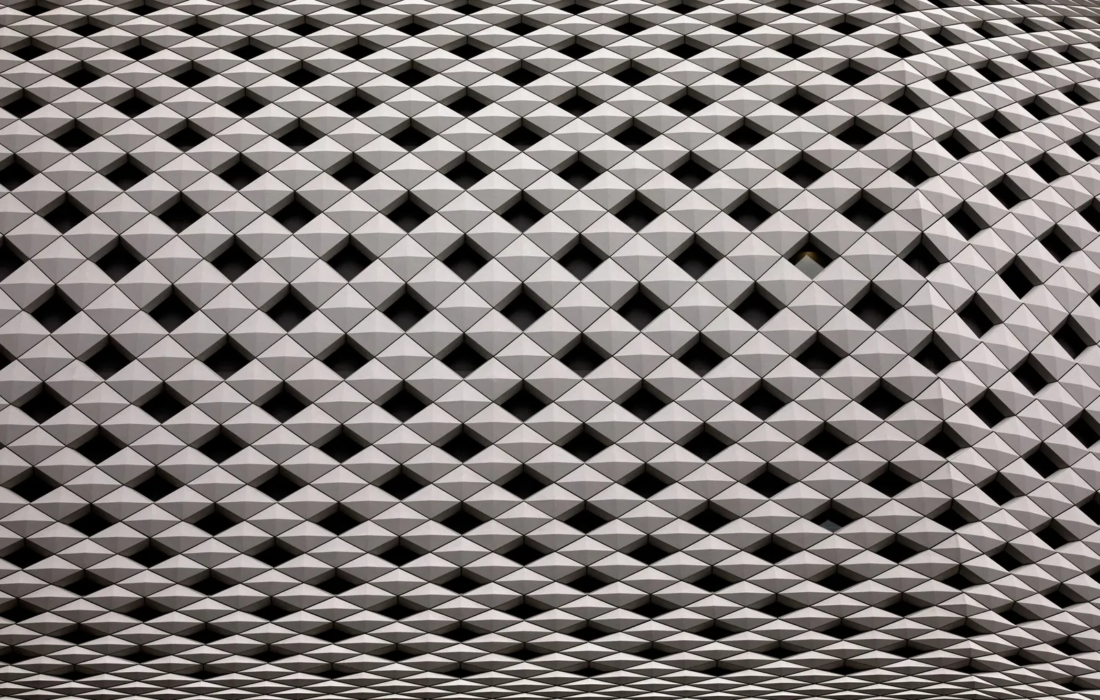 close view of a building surface covered in a pyramid pattern of pale gray tiles and dark square openings, filling the frame