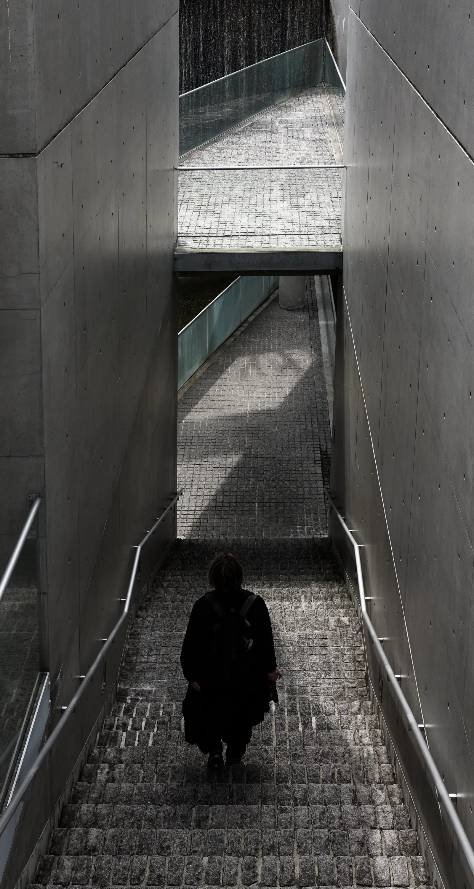 a person in dark clothing walking down a stone-paved stairway between tall concrete walls with metal handrails