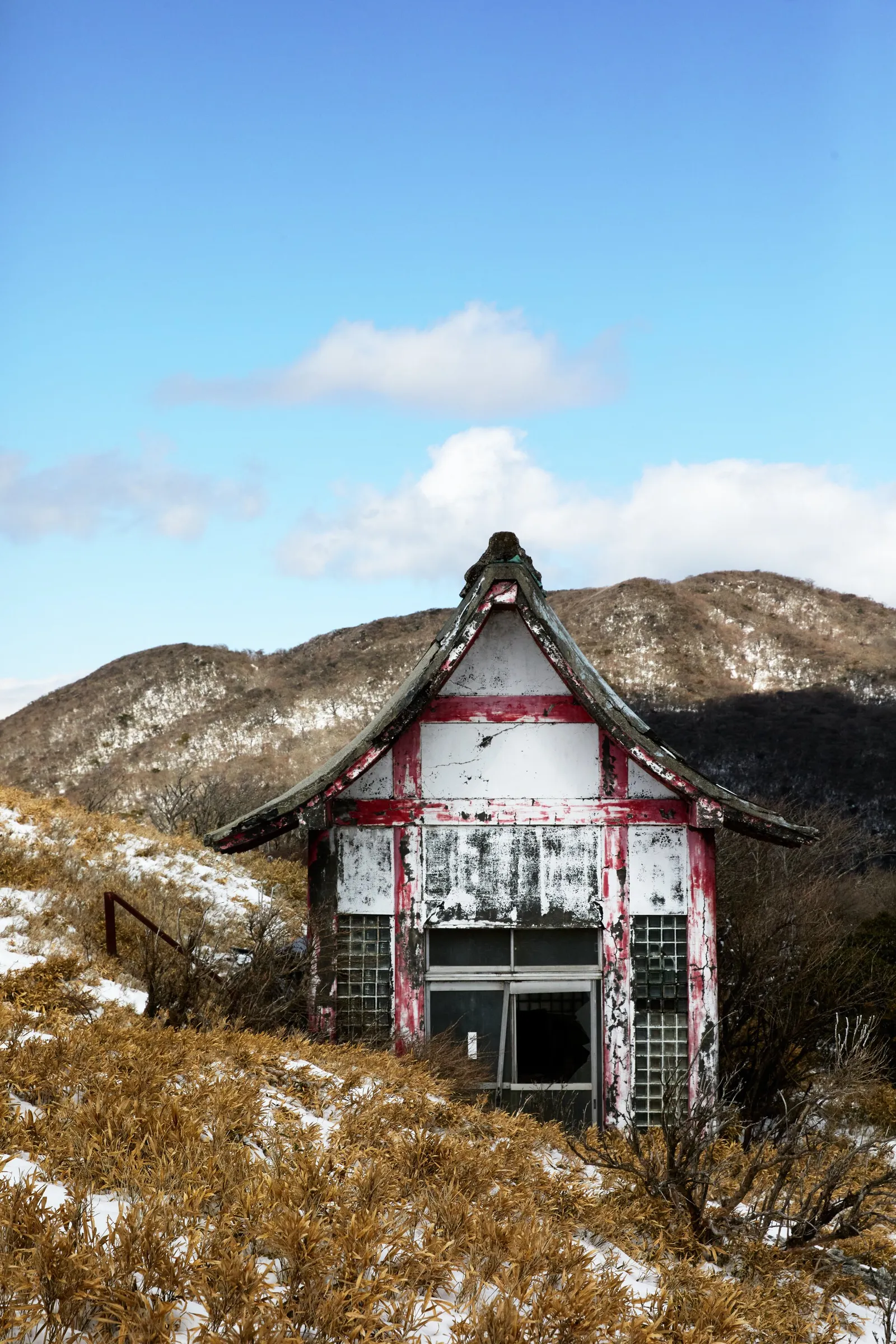 small abandoned shrine-like hut with peeling white and red paint on a snowy mountain slope of dry golden grass, blue sky behind