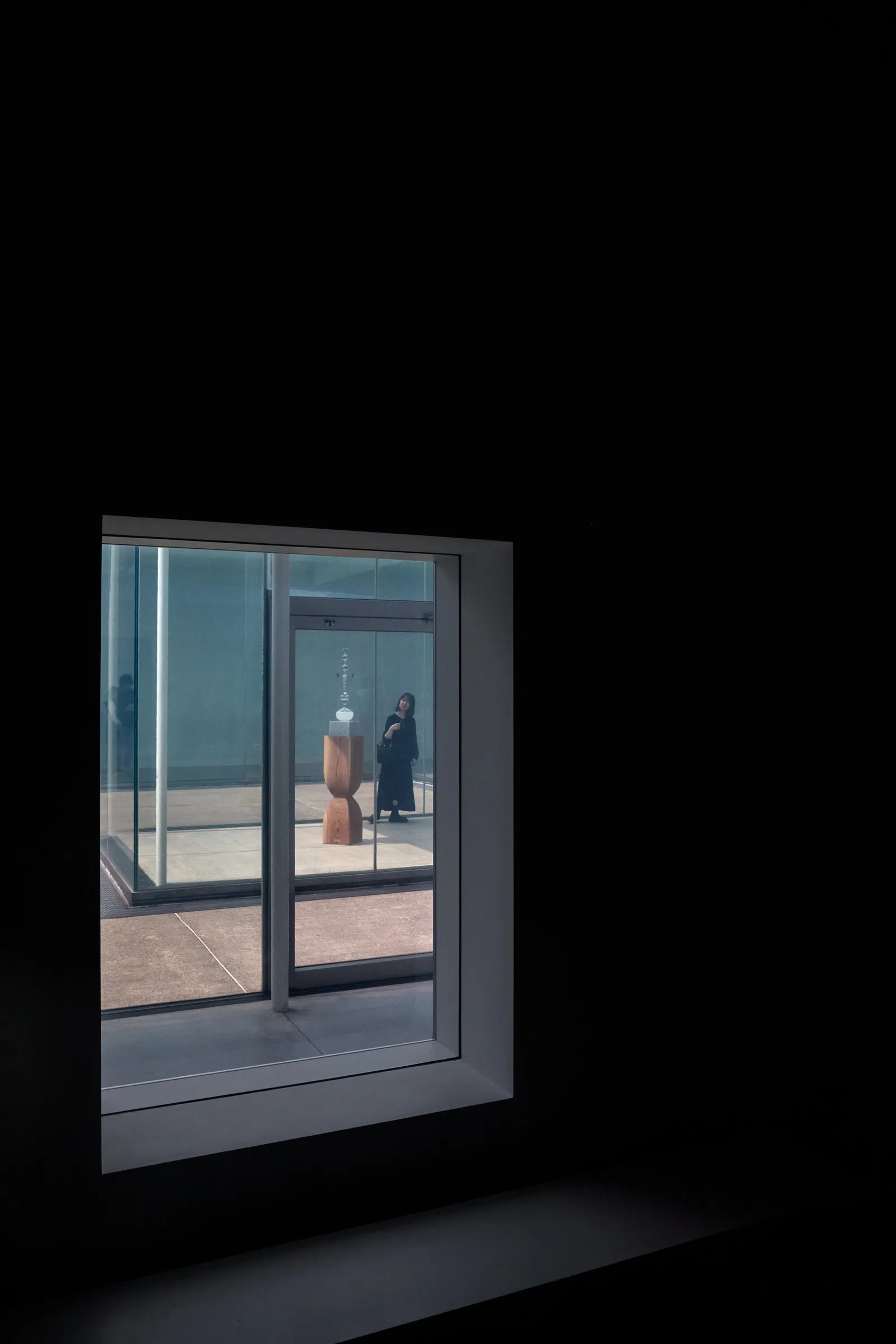 a lit window in a dark wall looking into a glass-walled room where a person stands near a tall wooden sculpture
